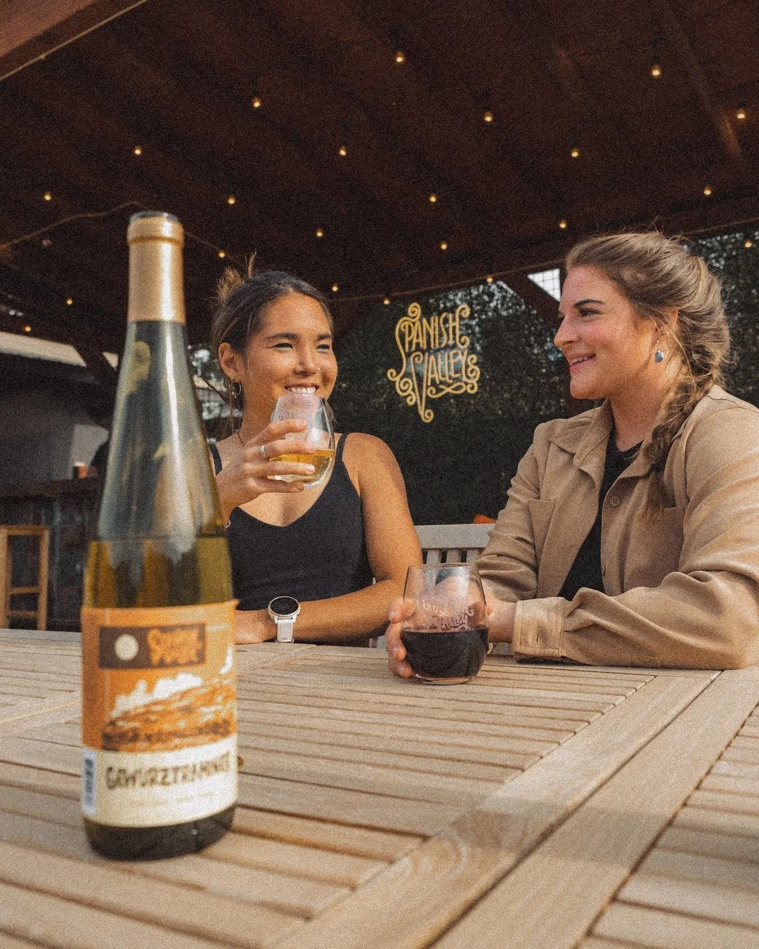 Two women sitting at a wooden table outside, smiling and talking, with a bottle of beer and two glasses of wine. The background has string lights and a sign that reads 'Pansic Alley'.