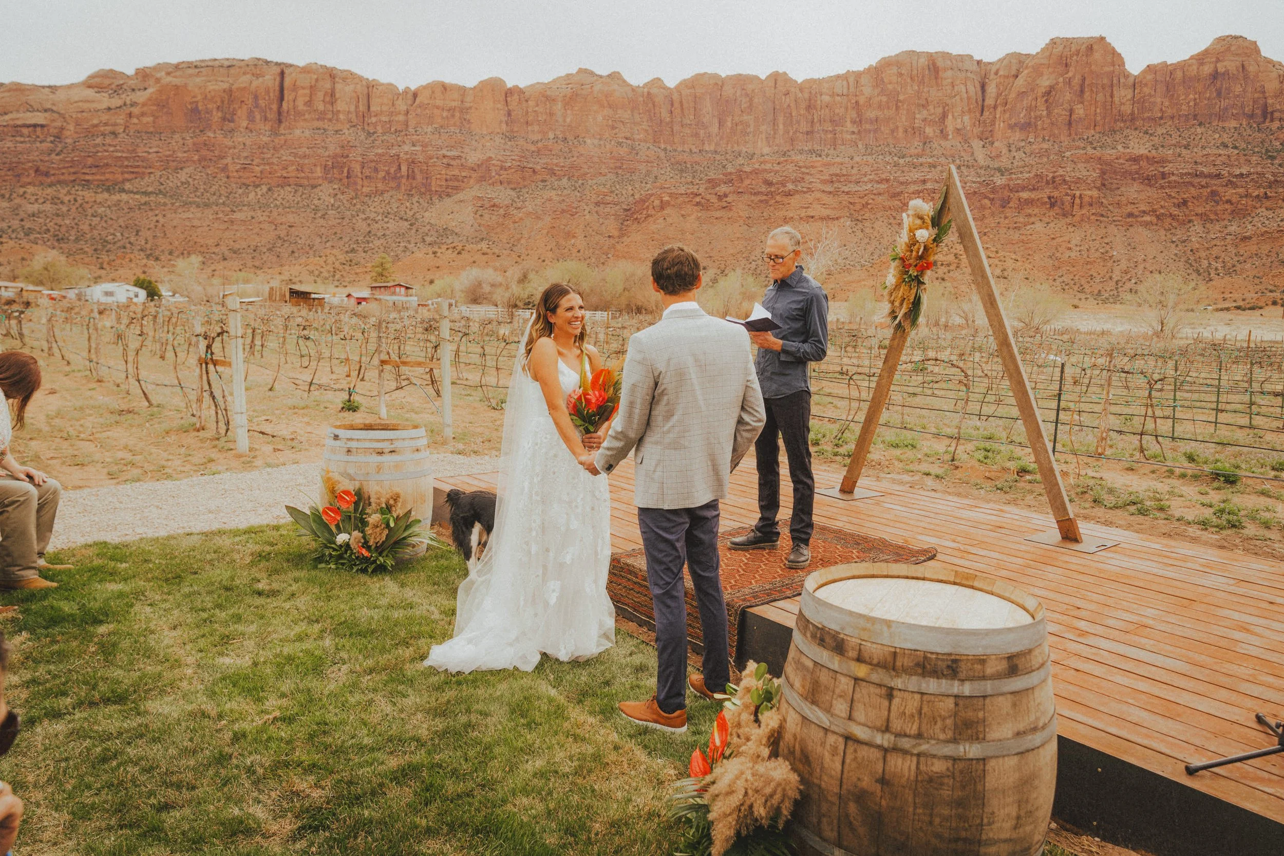 A couple getting married outdoors in a vineyard with red rock formations in the background, with a officiant, wedding arch, and floral decorations.