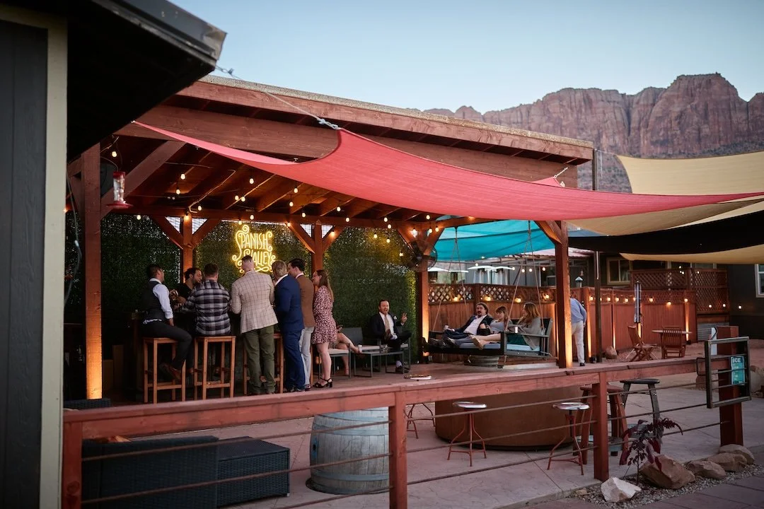 People socializing at an outdoor patio with string lights, wooden furniture, and a neon sign that reads 'Spanish Valley,' with mountains in the background.