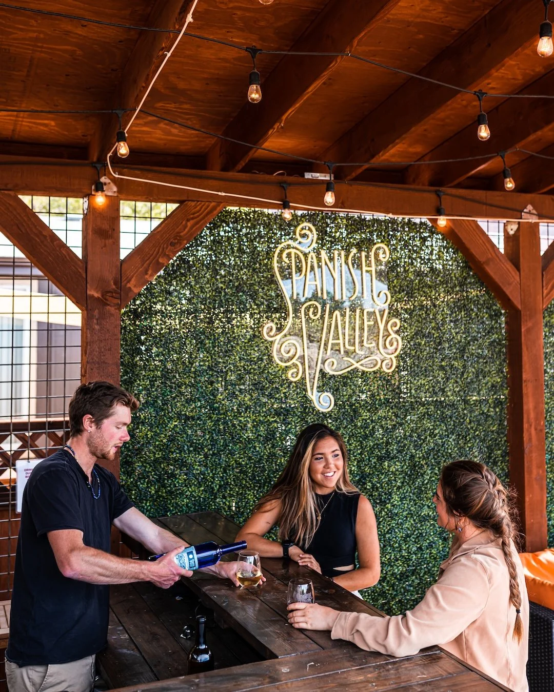 Three people at a bar with a green hedge background and a neon sign that says 'Spanish Alley'. A man is pouring wine into a glass for two women, who are smiling and sitting on the bar counter.