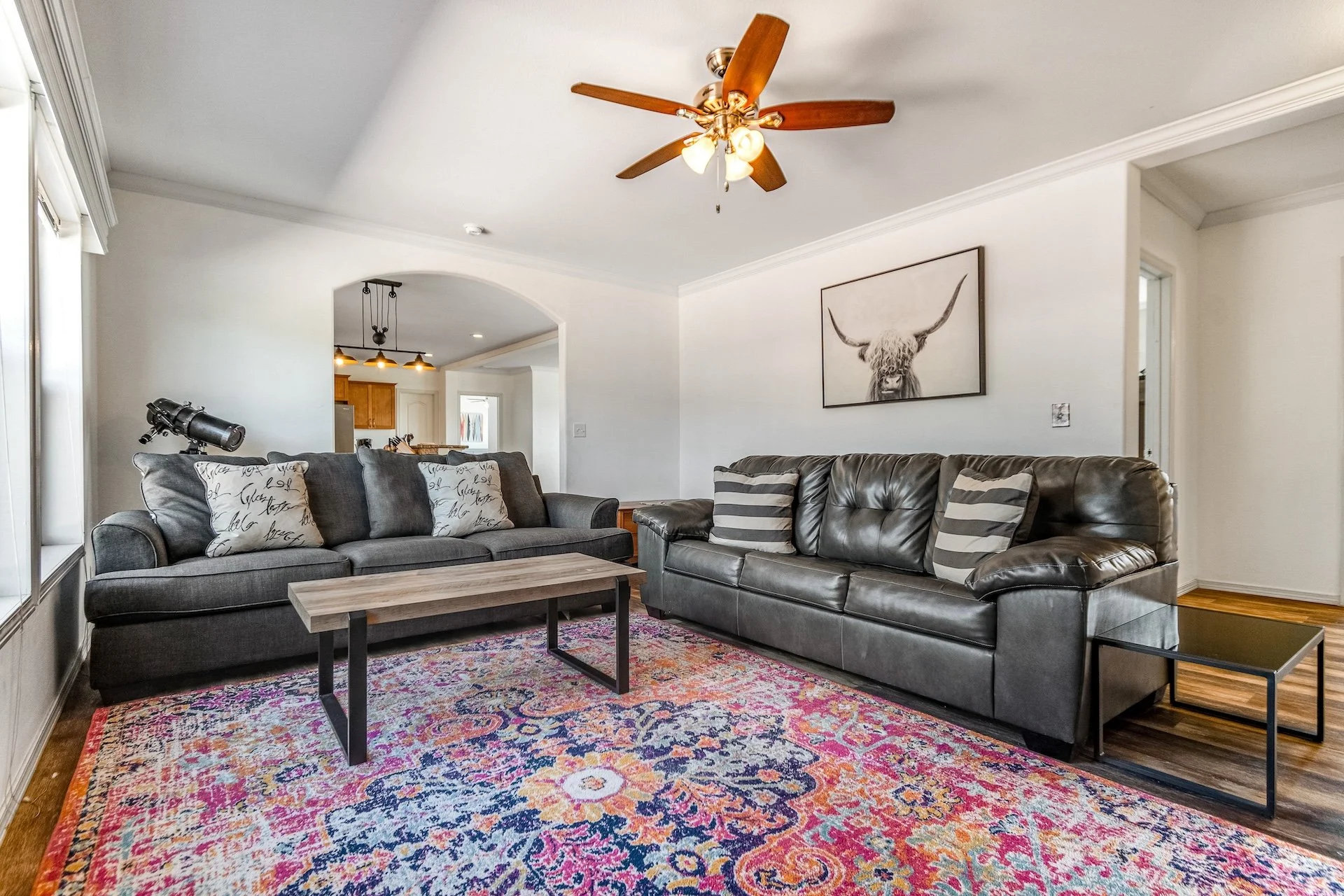 Living room with two gray sofas, a colorful patterned rug, a wooden coffee table, decorative pillows, a wall art of a highland cow, and a ceiling fan with wooden blades.
