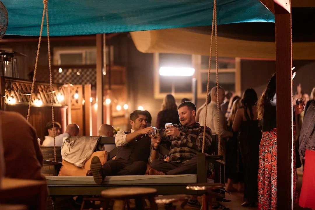 Two men sitting on a sofa, holding drinks, smiling and toasting at a lively indoor party or bar with other people in the background.