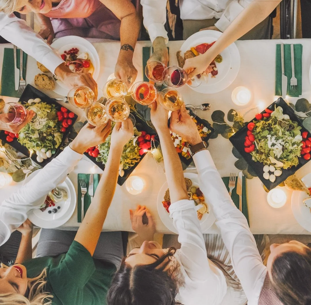 People raising glasses of wine in a toast over a dinner table with salads, candles, and festive decorations.