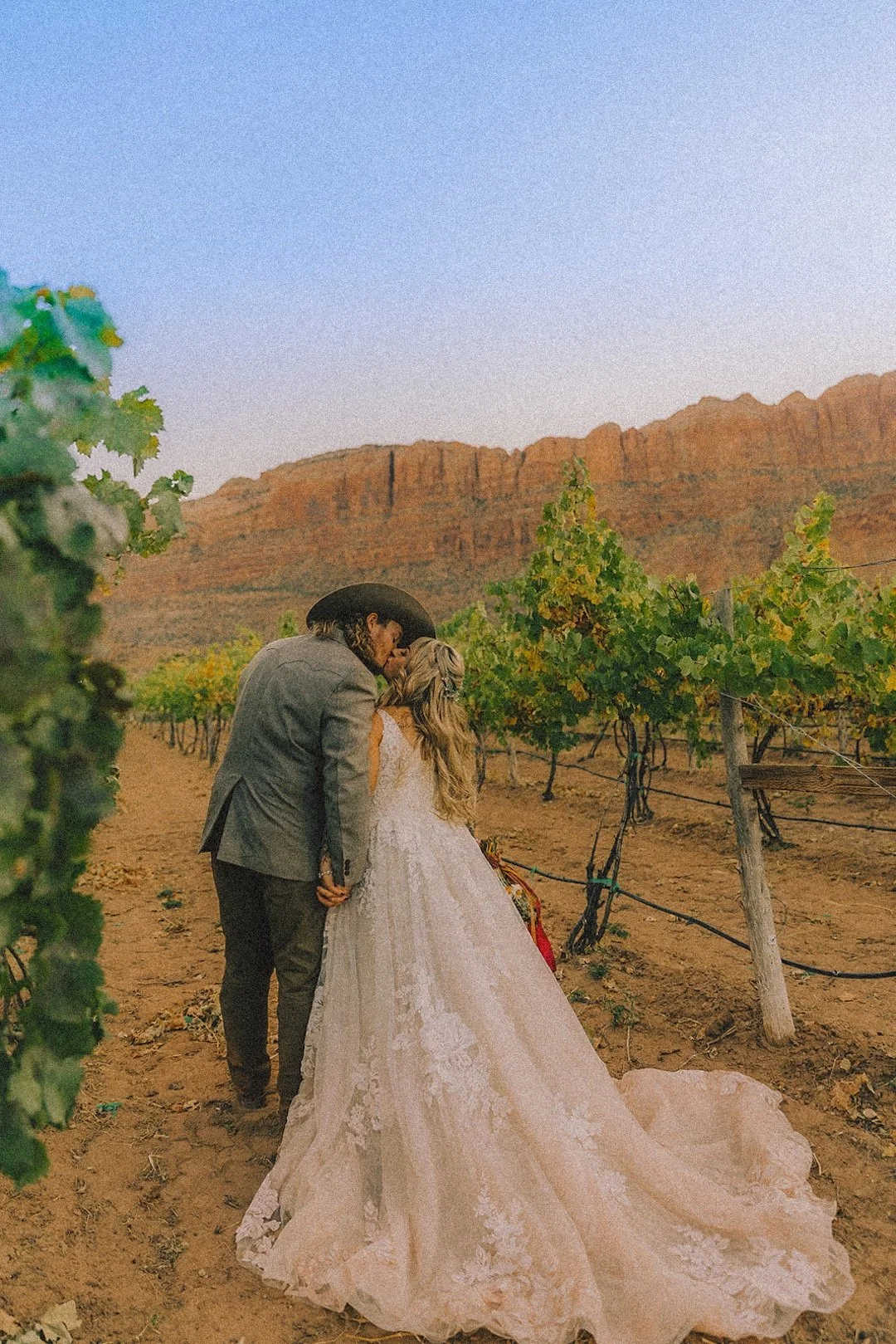 A couple dressed in wedding attire sharing a kiss in a vineyard with a red rock canyon in the background.