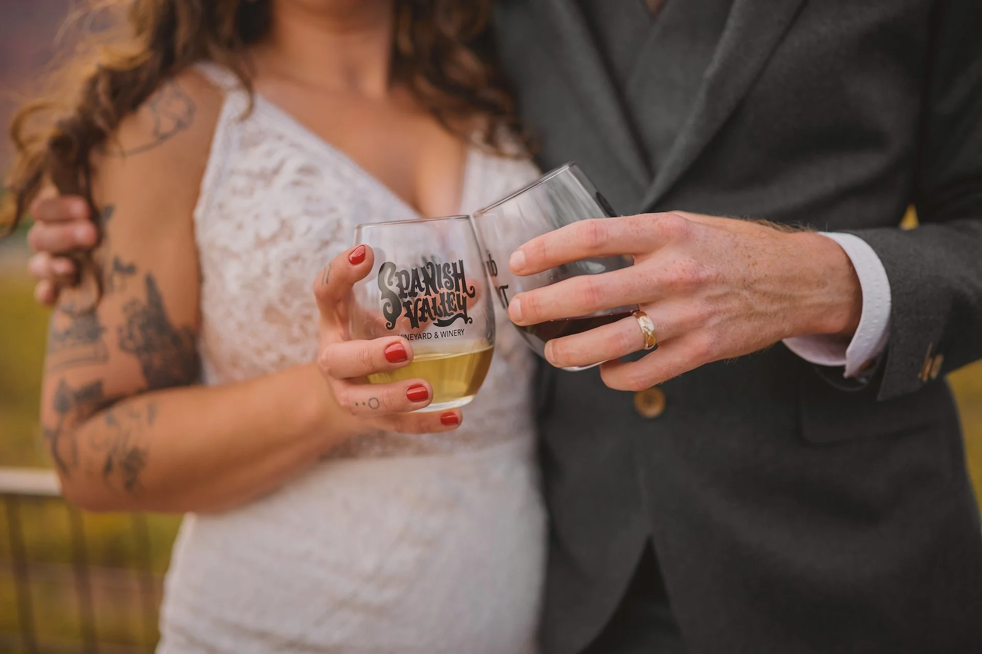Close-up of a couple holding wine glasses with a vineyard logo, a woman with tattoos and red nails wearing a lace dress, and a man in a gray suit, sharing a toast outdoors.