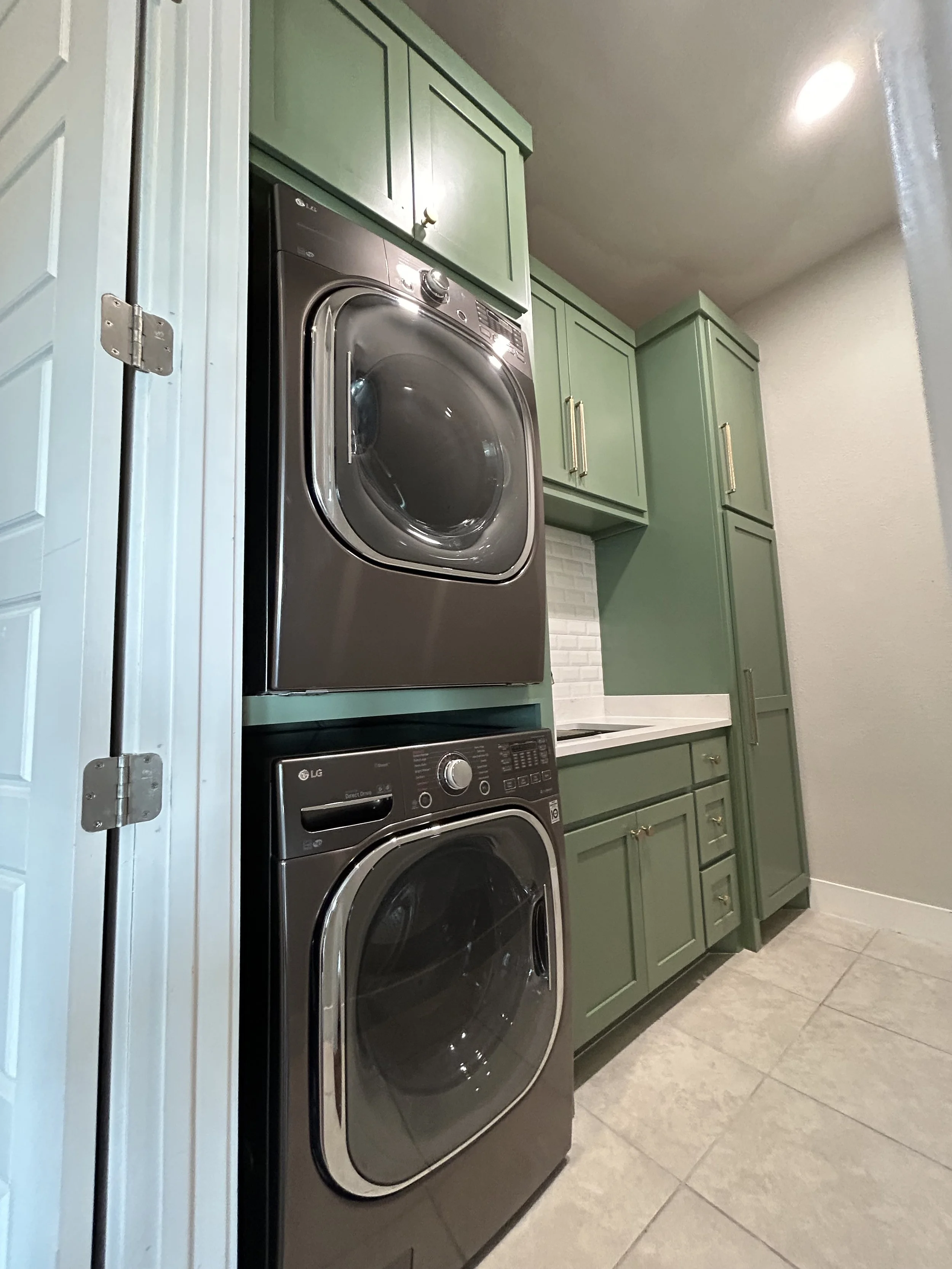 Stacked black front-loading washer and dryer in a laundry room with green cabinets and white backsplash.