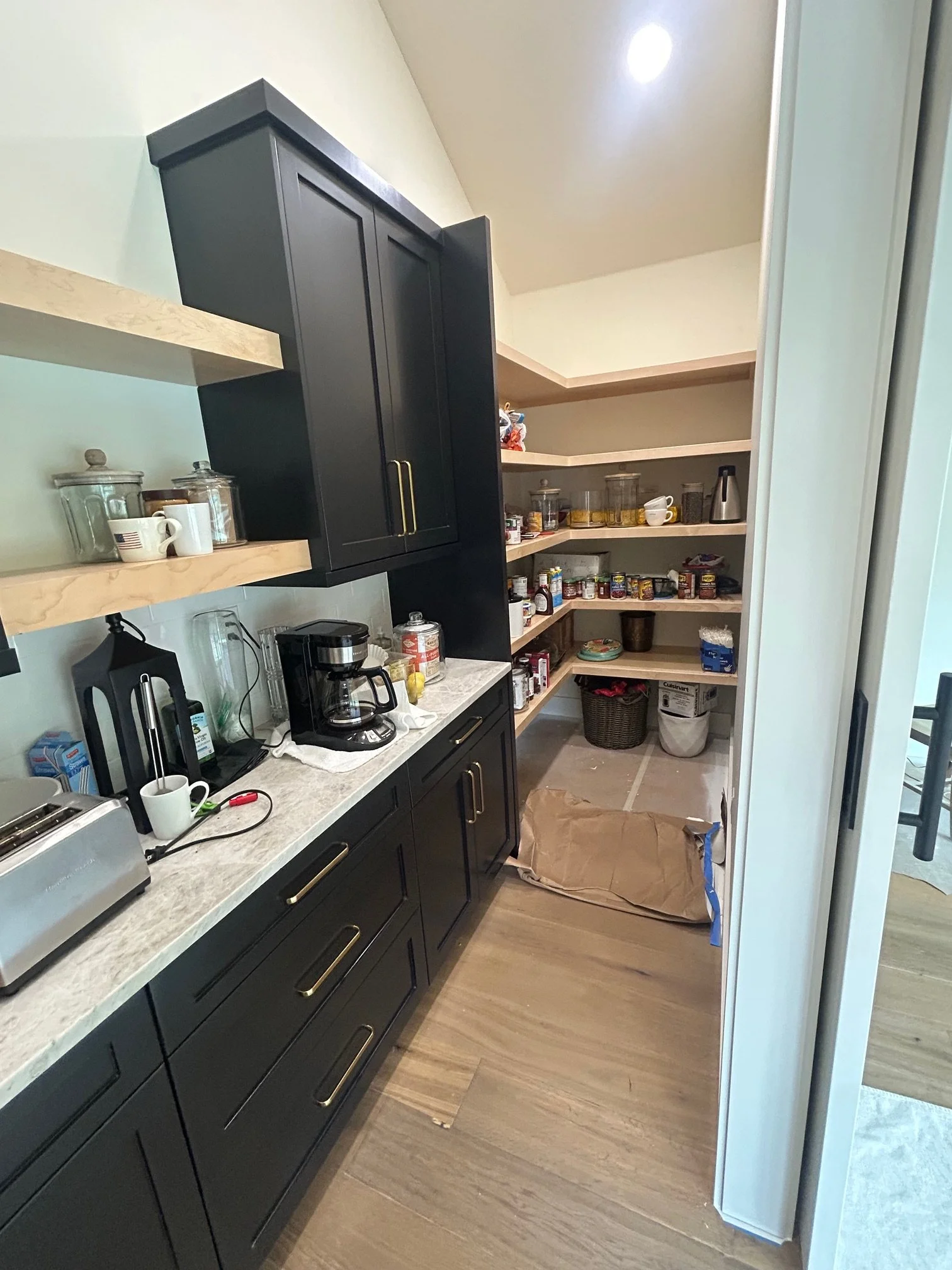 Pantry with open shelves filled with jars, canned goods, and food items, adjacent to a kitchen counter with coffee maker, toaster, and cups.