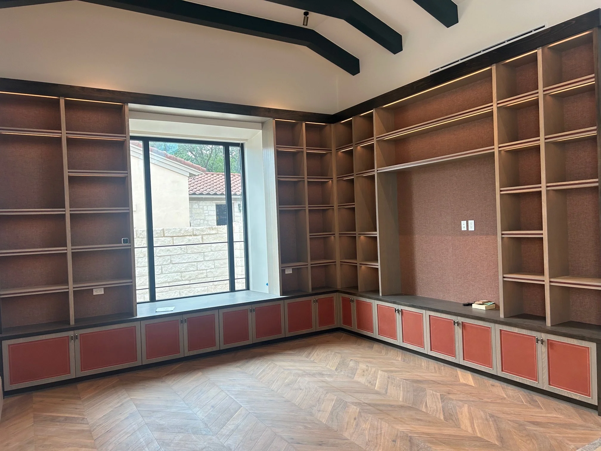 Empty built-in wooden bookshelves with open shelves and closed cabinets below, inside a room with a large window.