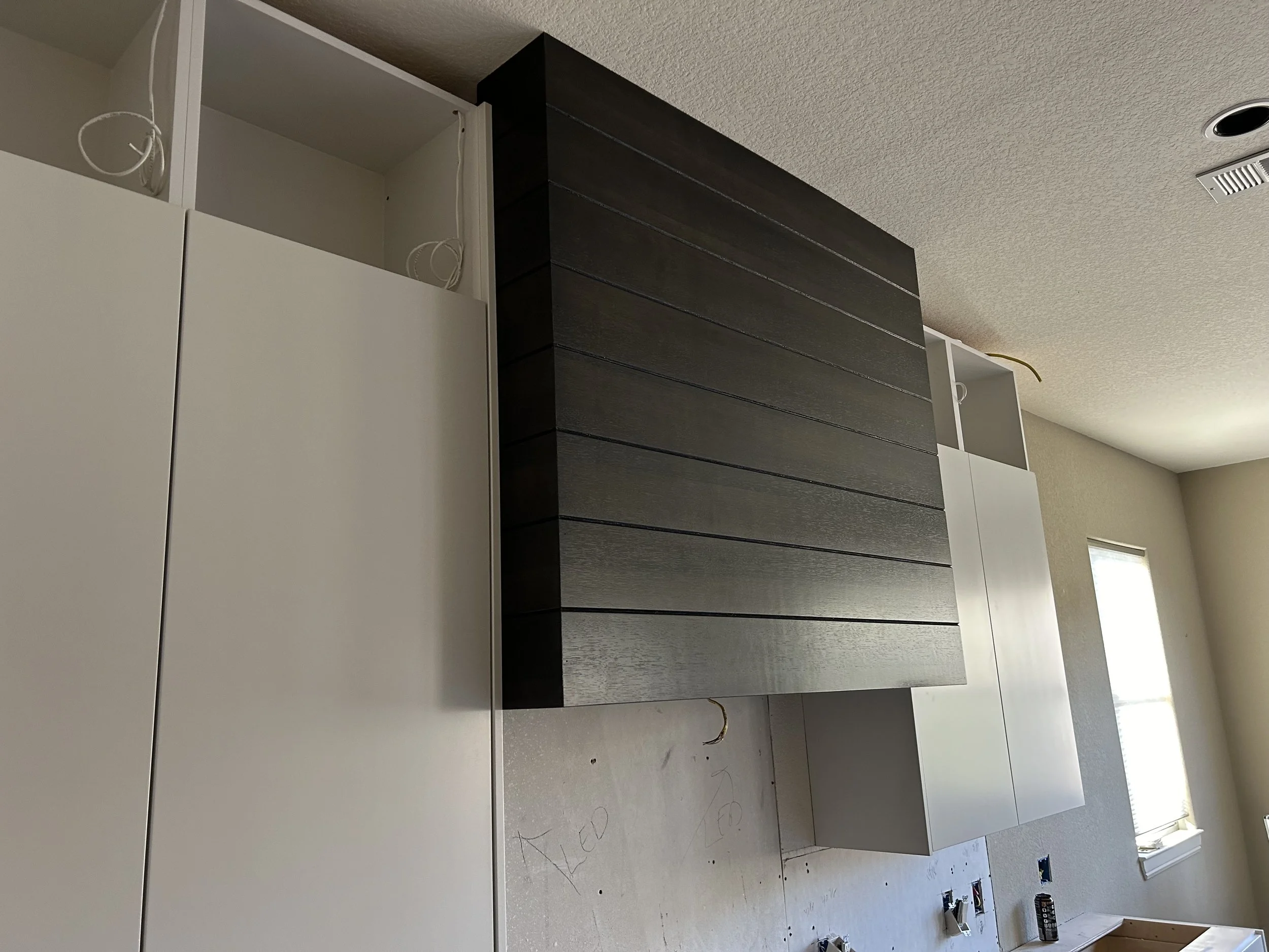 A room under construction with white cabinets and a black wooden panel attached to the wall. There are exposed wires and construction tools on the floor, and a window providing natural light.