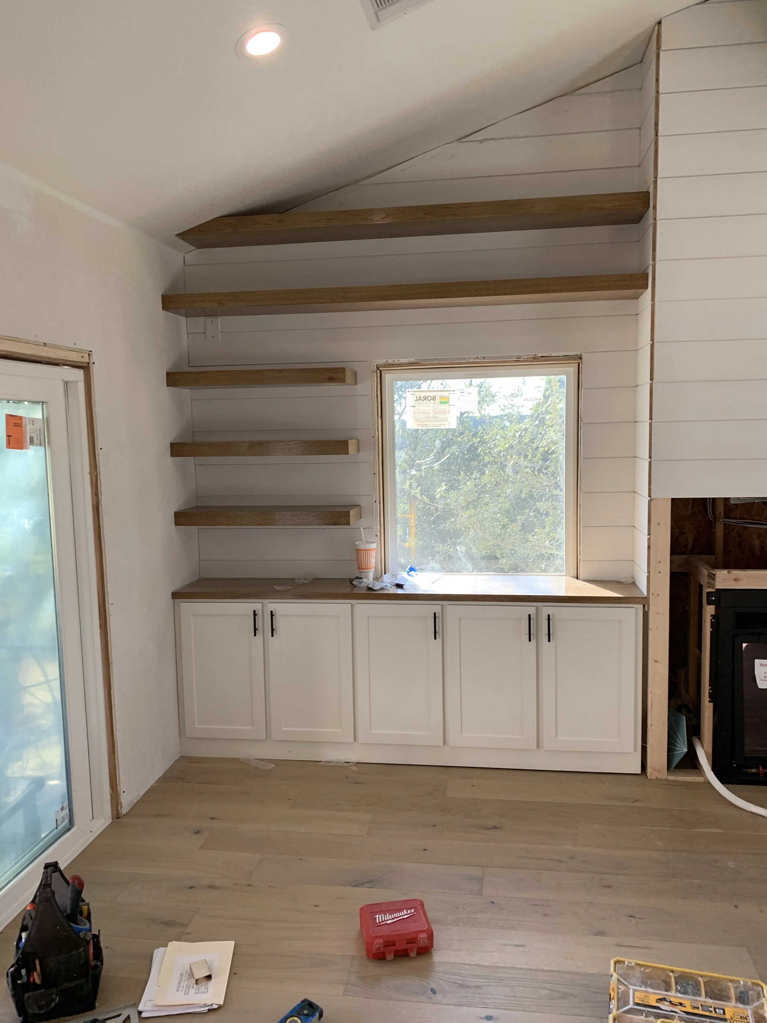 Interior of a room under construction with built-in white cabinets and open shelves above, a window letting in natural light, and construction tools on the floor.