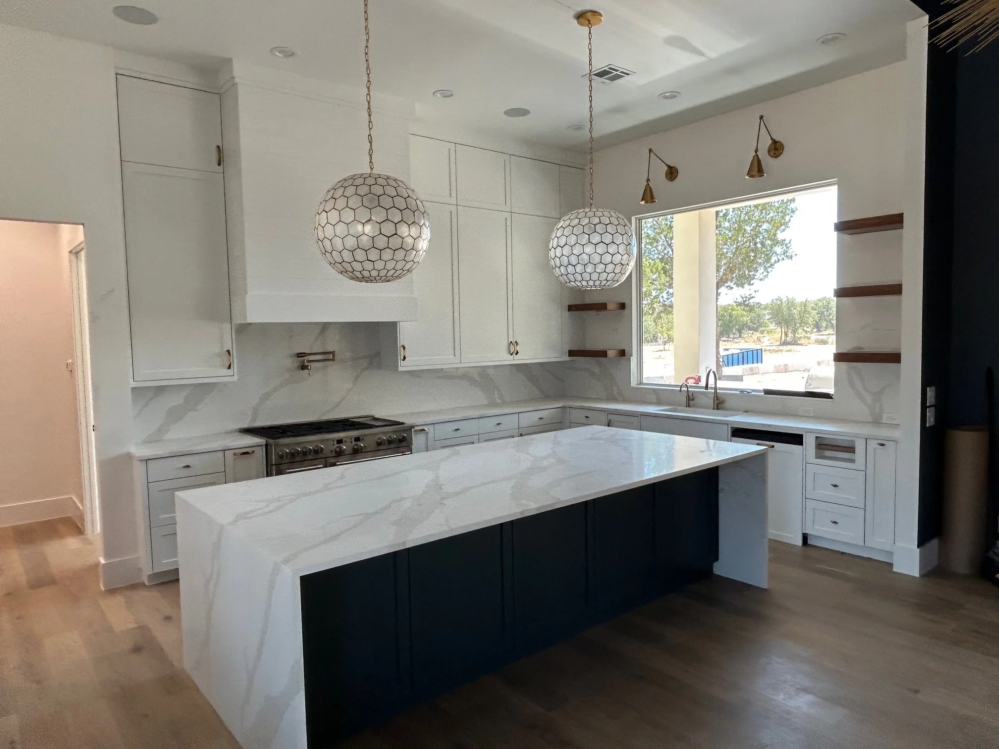 Modern kitchen with white cabinets and marble countertops, black lower cabinets on the island, and two large hanging pendant lights with honeycomb patterns. There are small spotlights and a large window overlooking a yard with trees.