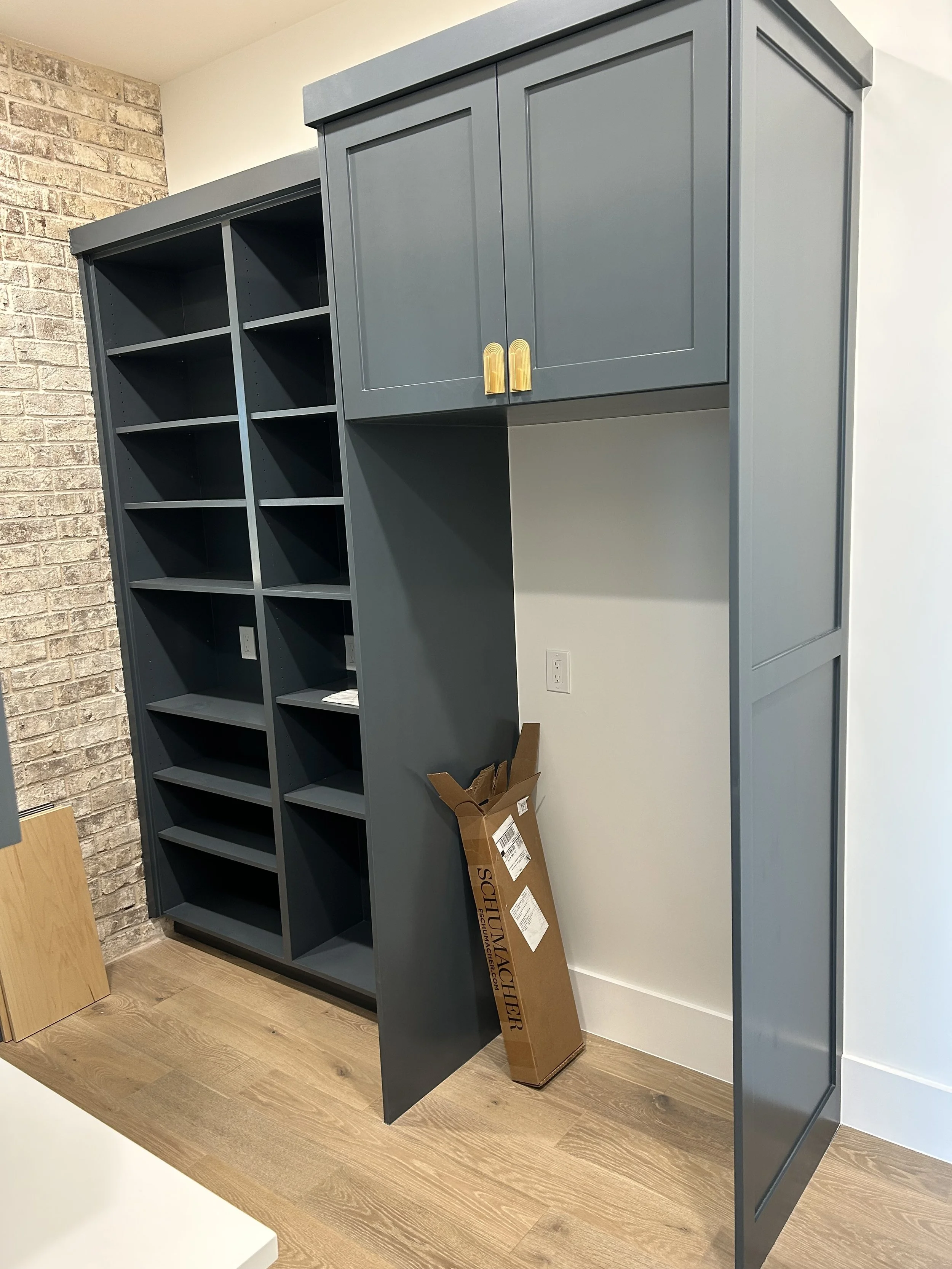 Blue built-in shelves and cabinets in a room, some empty, with a brick wall on the left, a cardboard box labeled 'Schluter' on the floor, and part of a white table in the foreground.