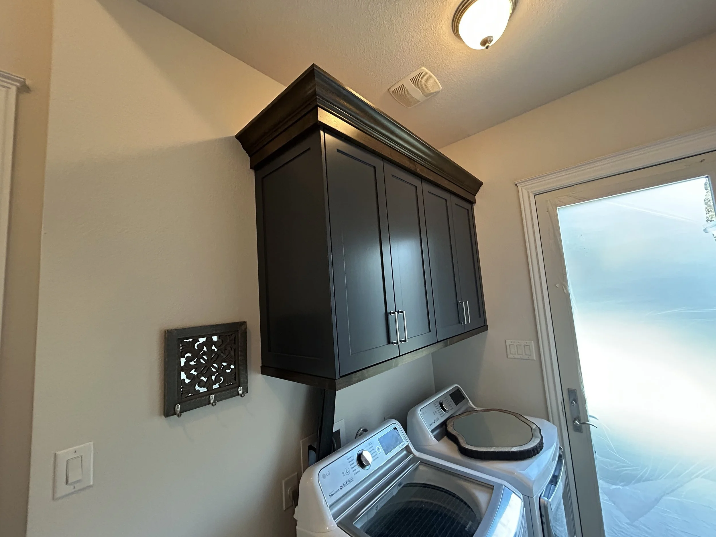 A laundry room with a white washer and dryer, dark wall-mounted cabinets above, a small framed decorative vent on the wall, a ceiling light fixture, and a glass door letting in natural light.