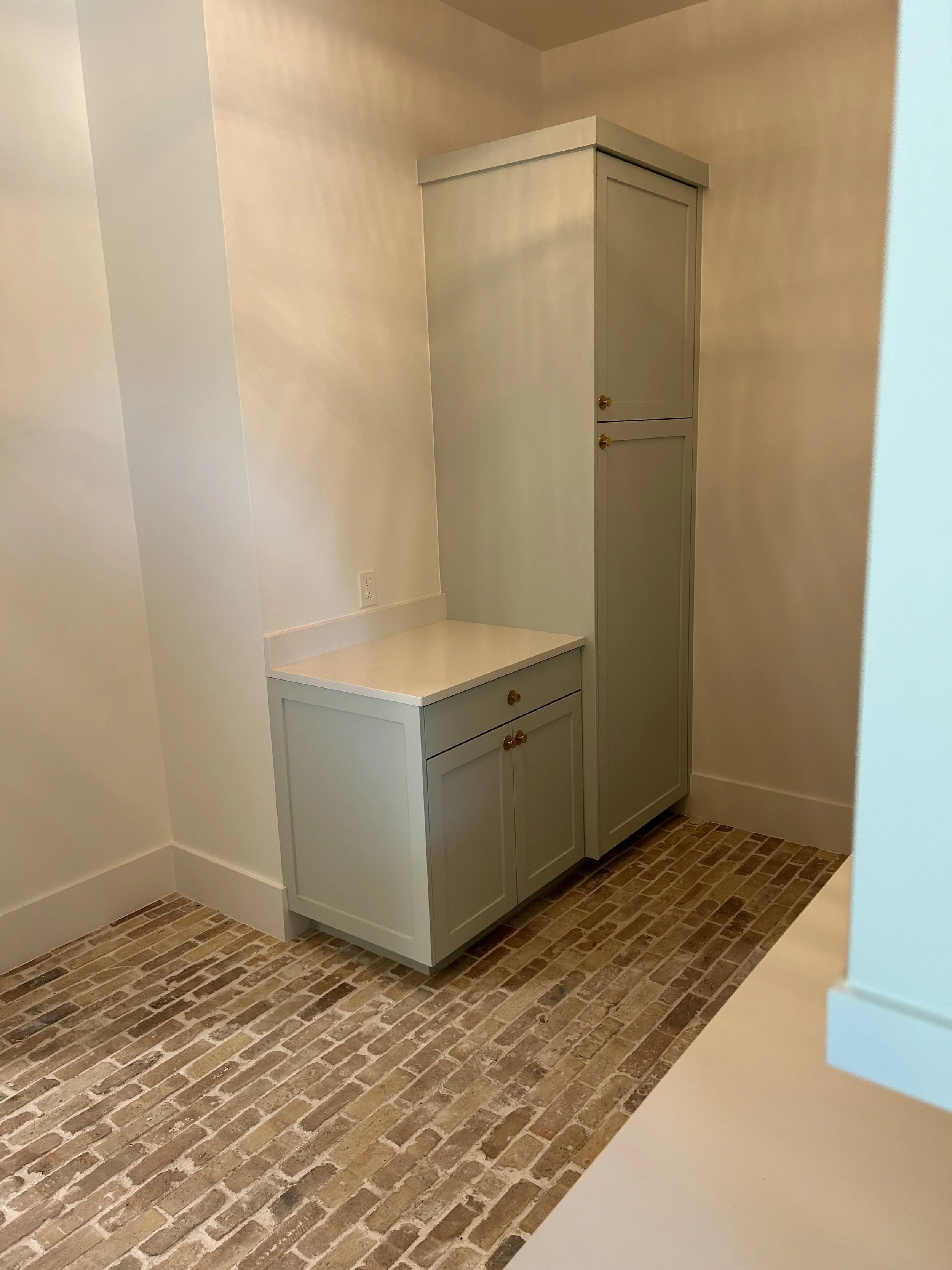 A corner of a room with beige walls and a brick-patterned floor. There is a white cabinet with gold knobs against the wall, comprising two sections: a smaller lower part and a taller upper part.