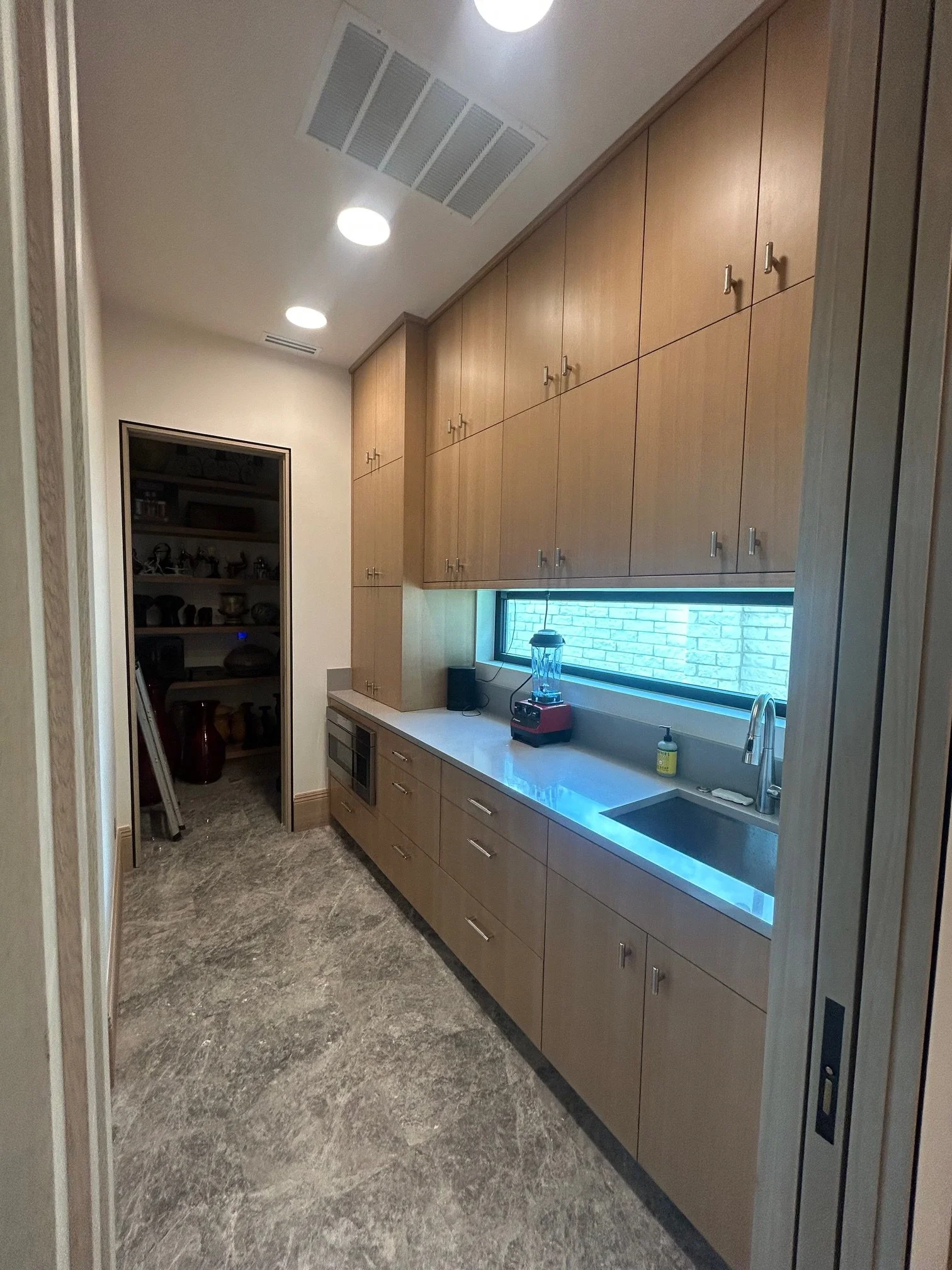A modern kitchen with wooden cabinets, a white countertop, a stainless steel sink, a blender, a microwave, and a window with a brick wall outside.