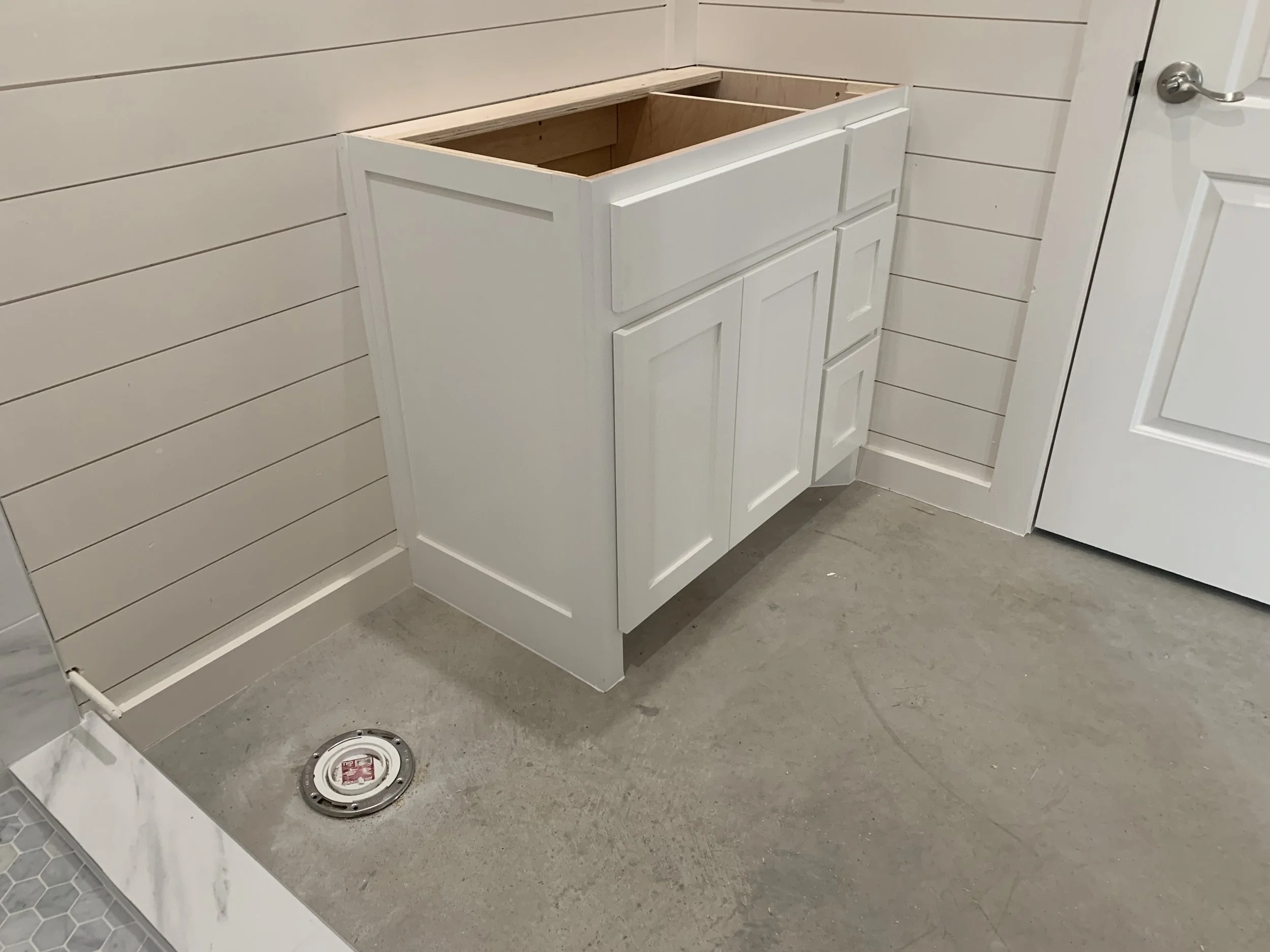 A bathroom with a white cabinet under construction, a wood-paneled wall, a drain in the concrete floor, and a door on the right side.