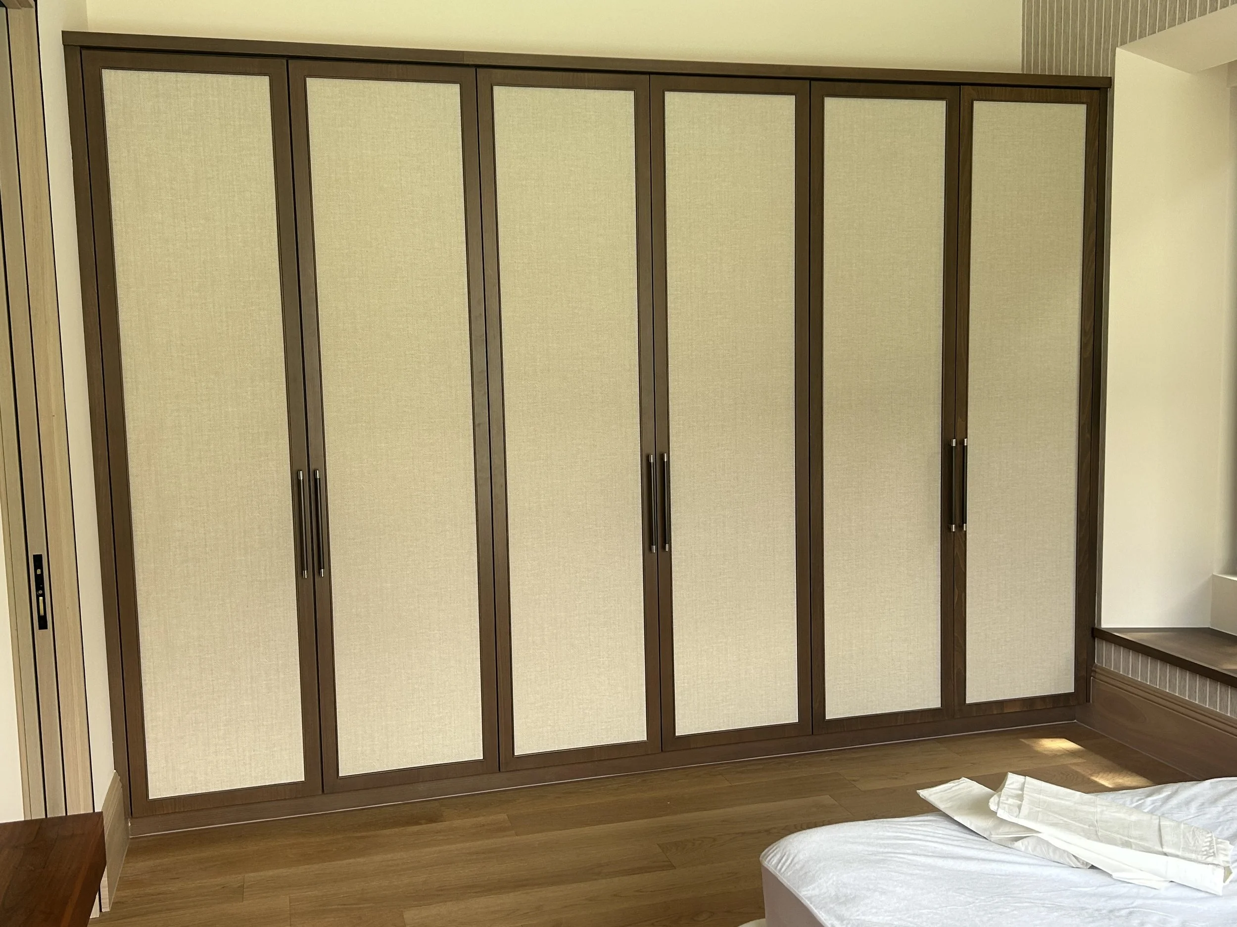 A wooden wardrobe with beige fabric-paneled doors in a room with wooden flooring and a small bedside table visible at the edge of the bed.