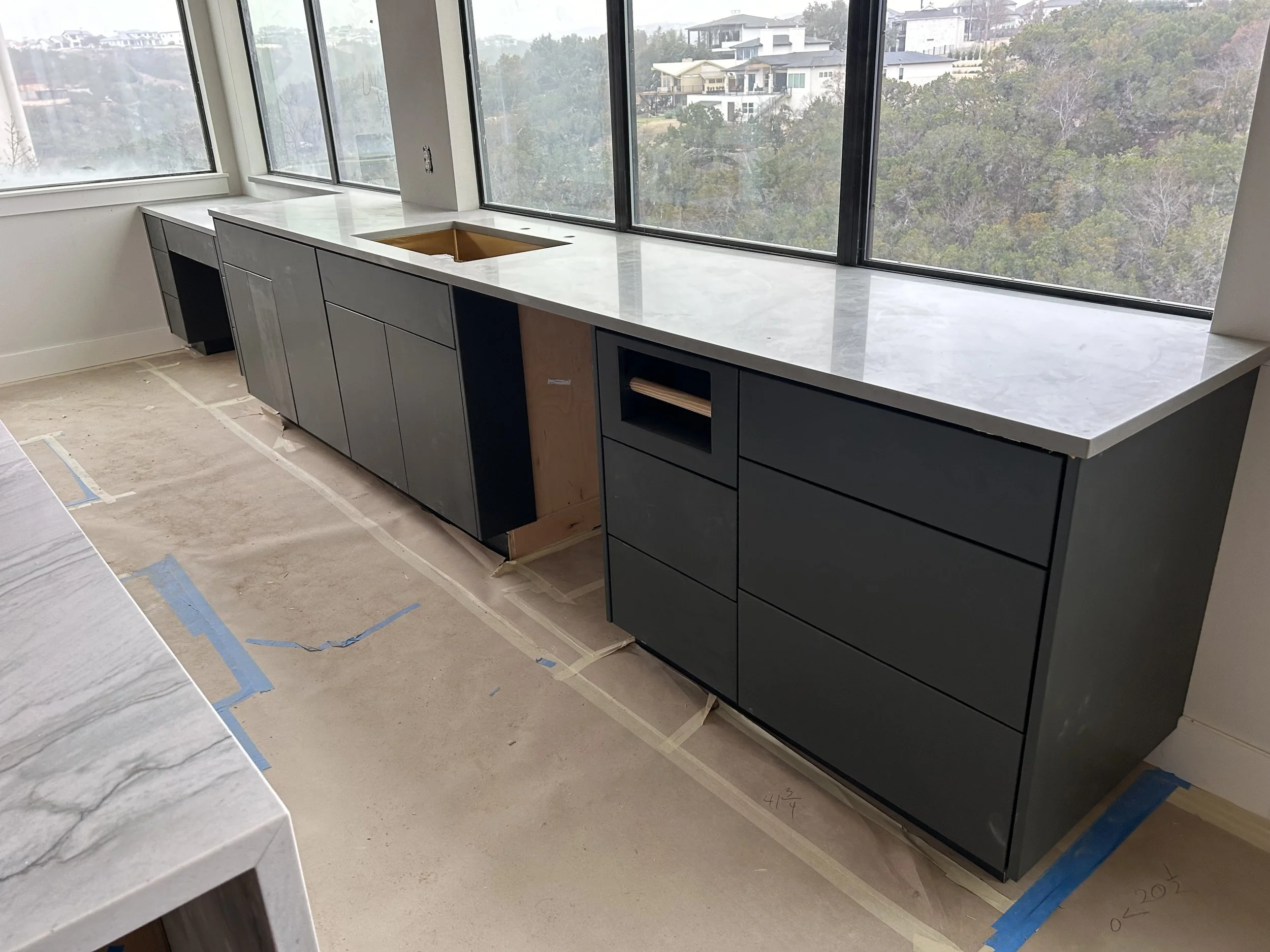 Kitchen cabinets under construction with a large marble countertop, a center cutout for a sink, and large windows overlooking a neighborhood with trees and houses.