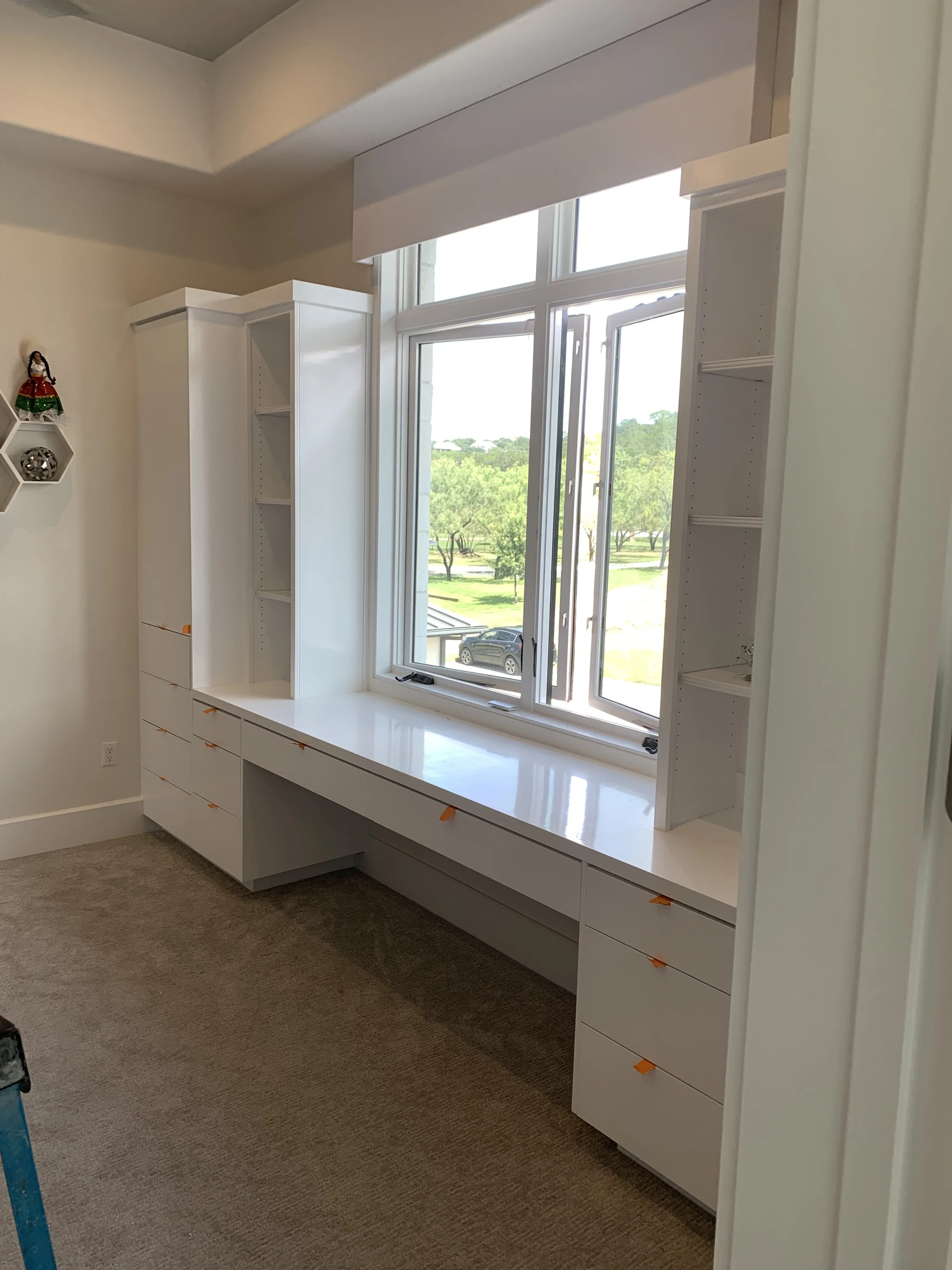 White custom-built window seat with multiple drawers and open shelves, large window with a view of trees, and a beige carpeted floor.