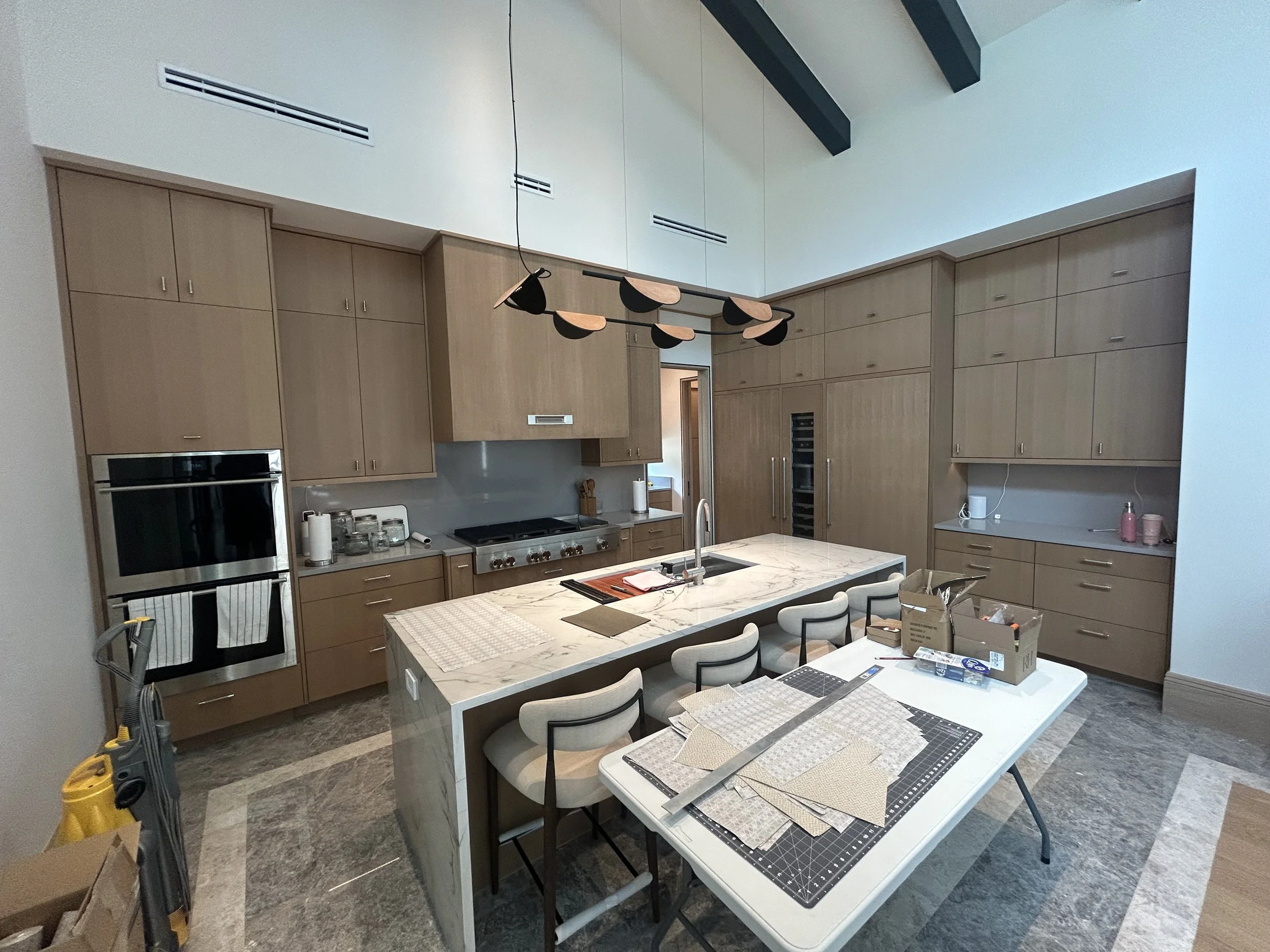 Modern kitchen with wooden cabinets, marble island, and ceiling beams. Items on the island include a sink, cutting board, and notebooks. A table with fabric samples, tools, and boxes is in the foreground, with a yellow vacuum on the left.