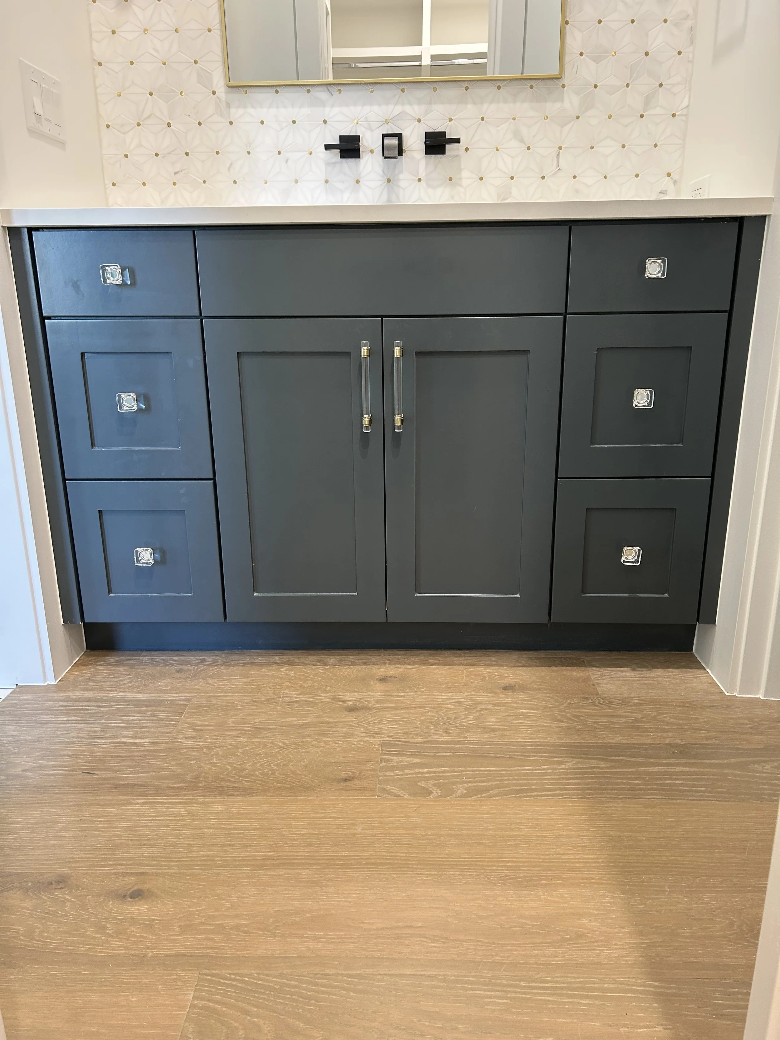 A bathroom vanity with dark gray cabinets and gold handles, set against a white wall with gold diamond pattern wallpaper, and a mirror above.