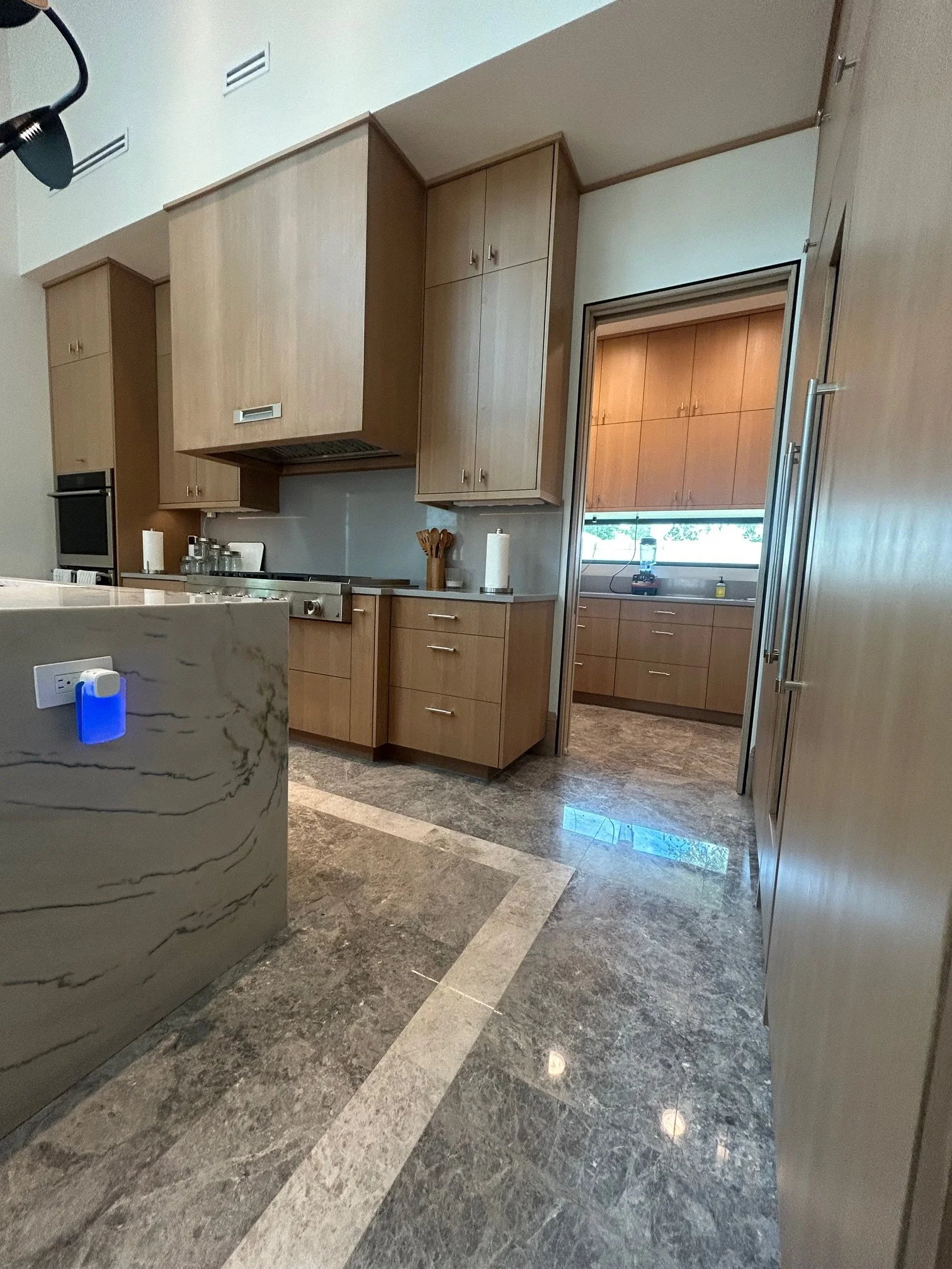 Modern kitchen with wooden cabinets, a marble countertop, and a polished stone floor. A doorway leads to a sunny room with similar cabinetry and window.