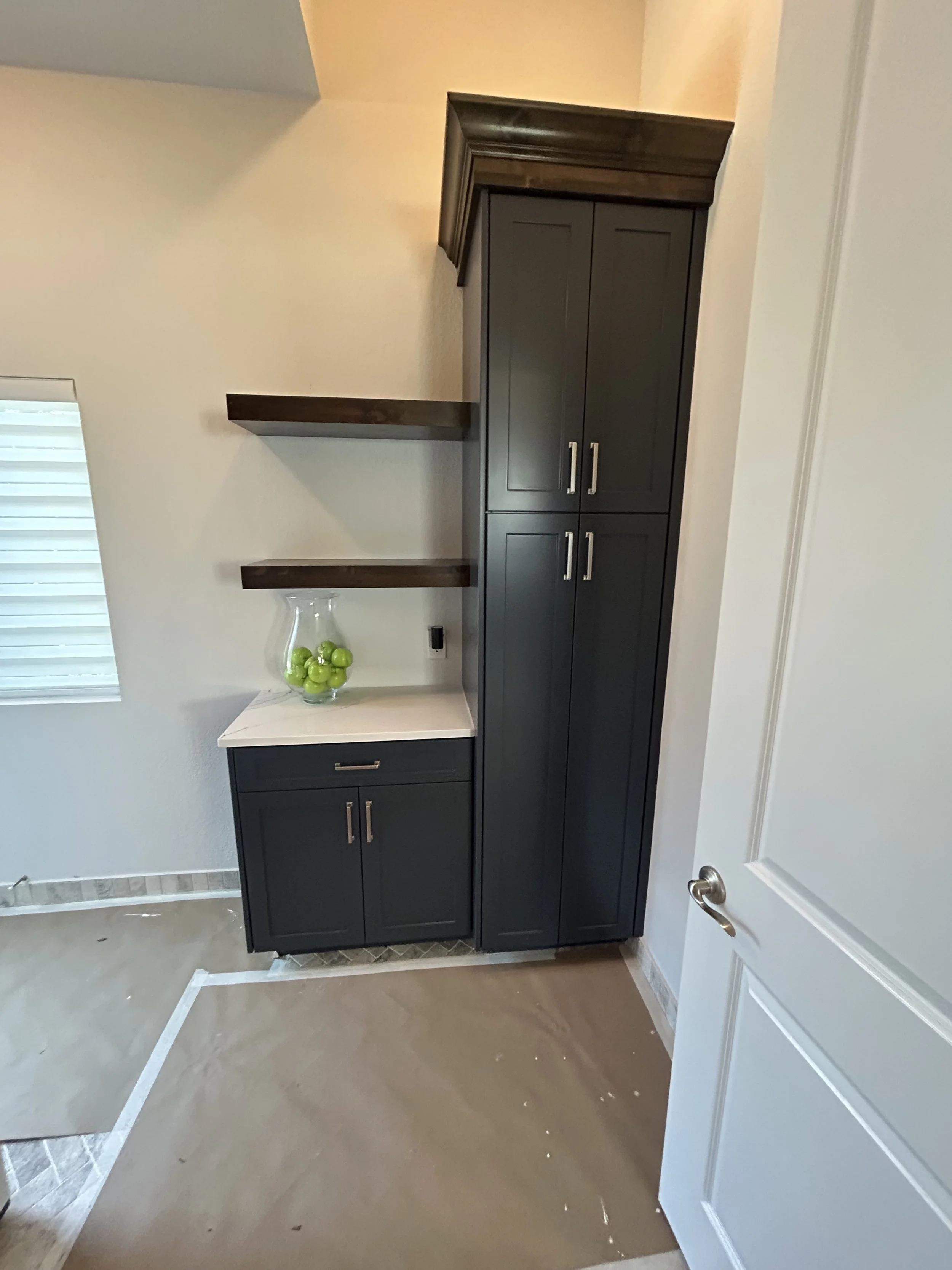 Corner of a room with dark cabinets, two open wooden shelves with a glass vase filled with green apples, and a window with closed blinds.