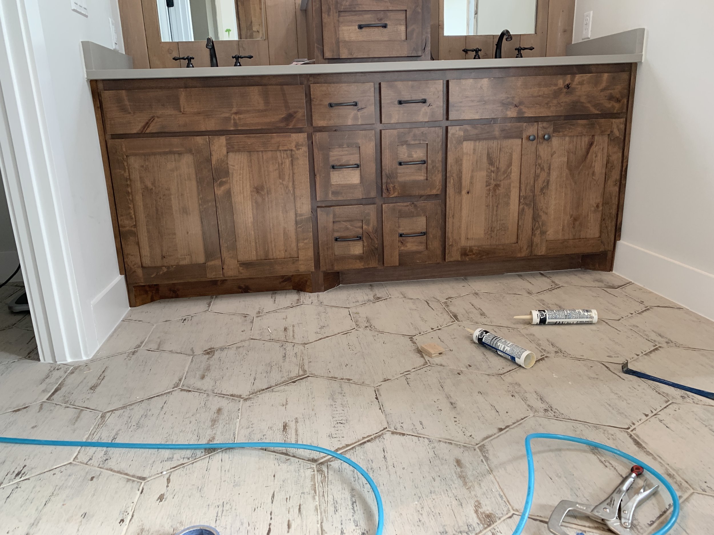 Bathroom vanity with a white countertop and black hardware. Construction tools and supplies on the floor, including caulk tubes, a small wooden piece, a paint scraper, and a blue hose.