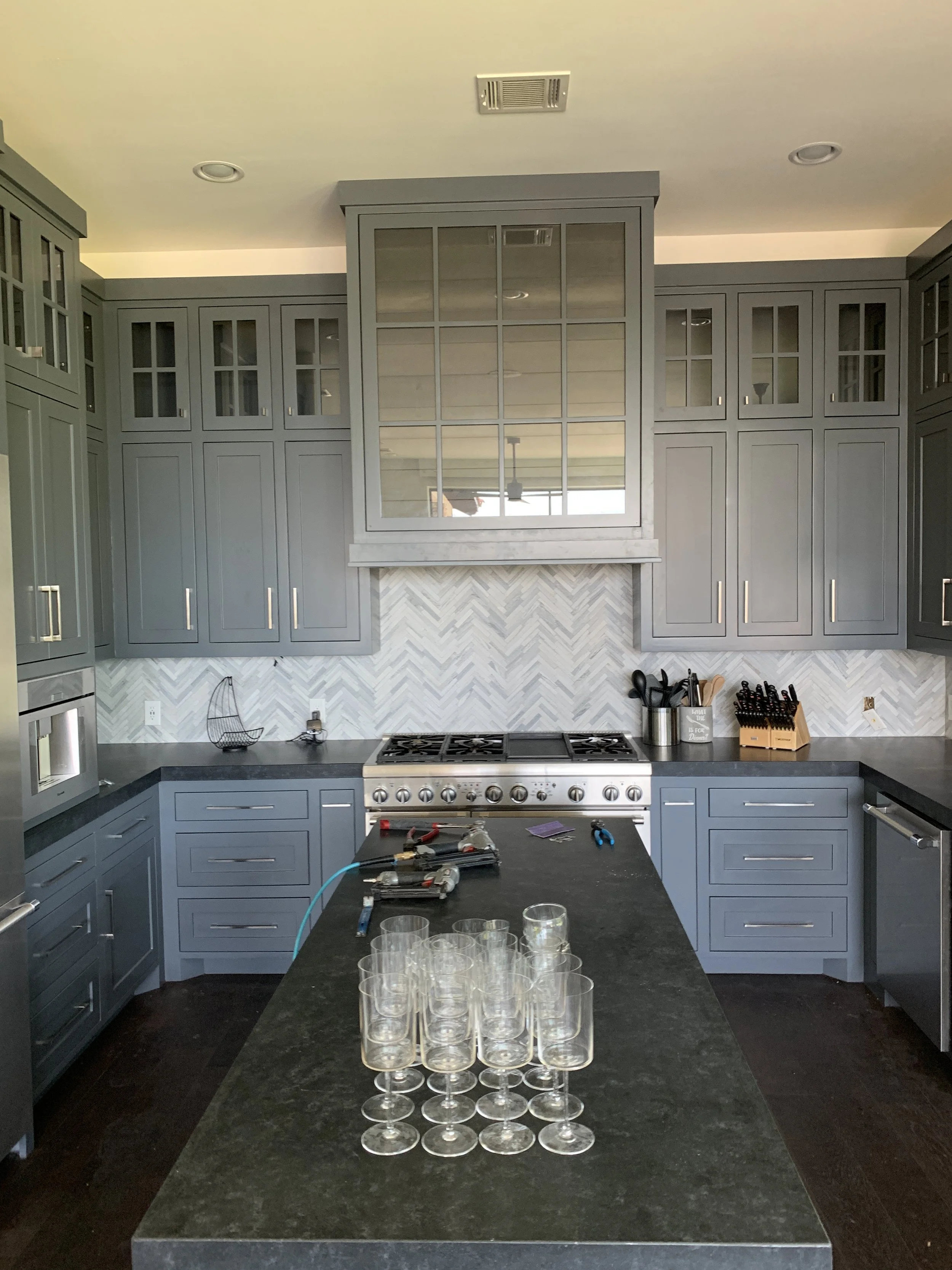 A modern kitchen with grey cabinets, black countertop, a gas stove, and glassware on the island counter.