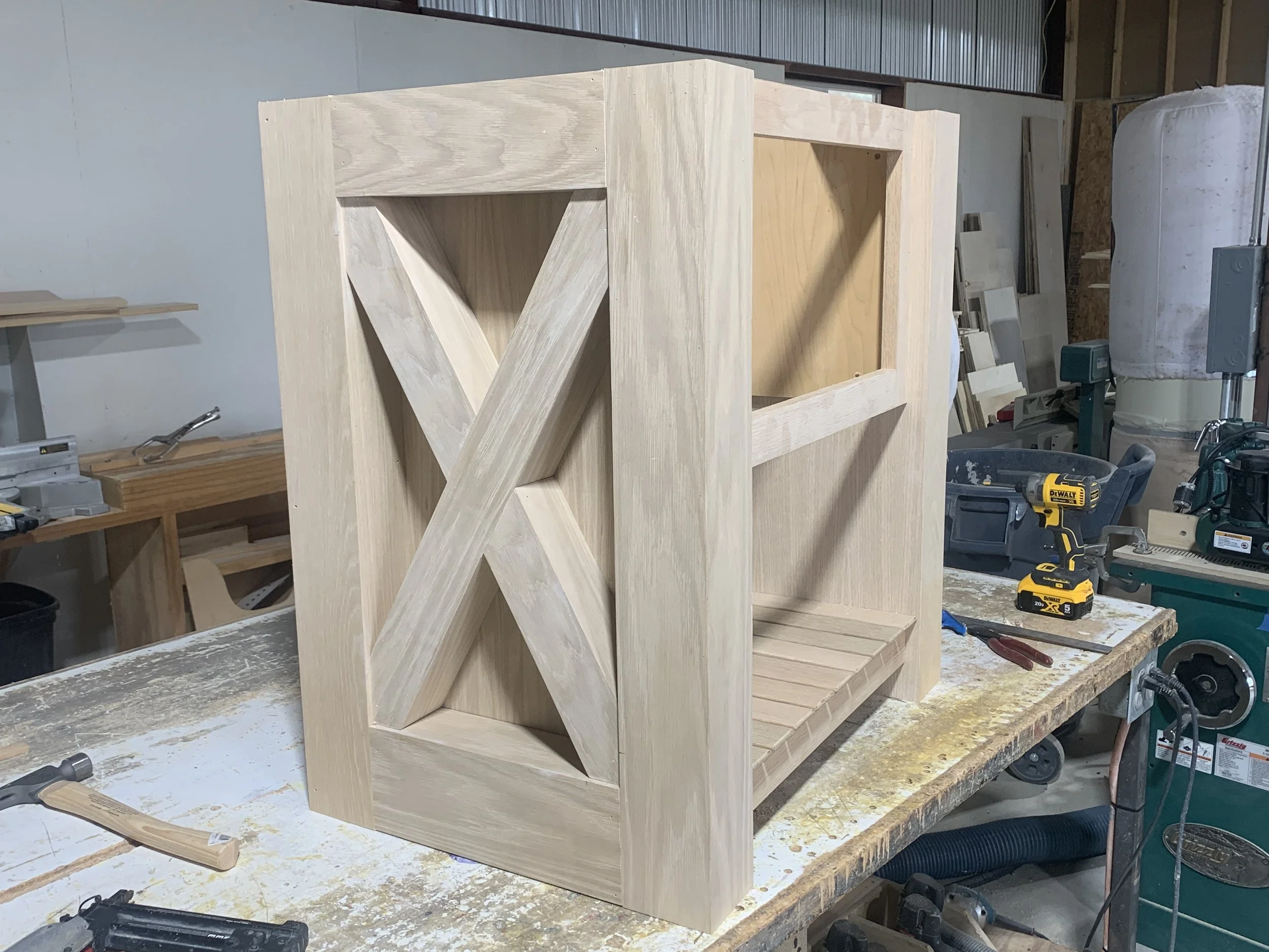 Wooden shelf or cabinet frame under construction on a workbench in a woodworking shop, with power tools and woodworking supplies around.
