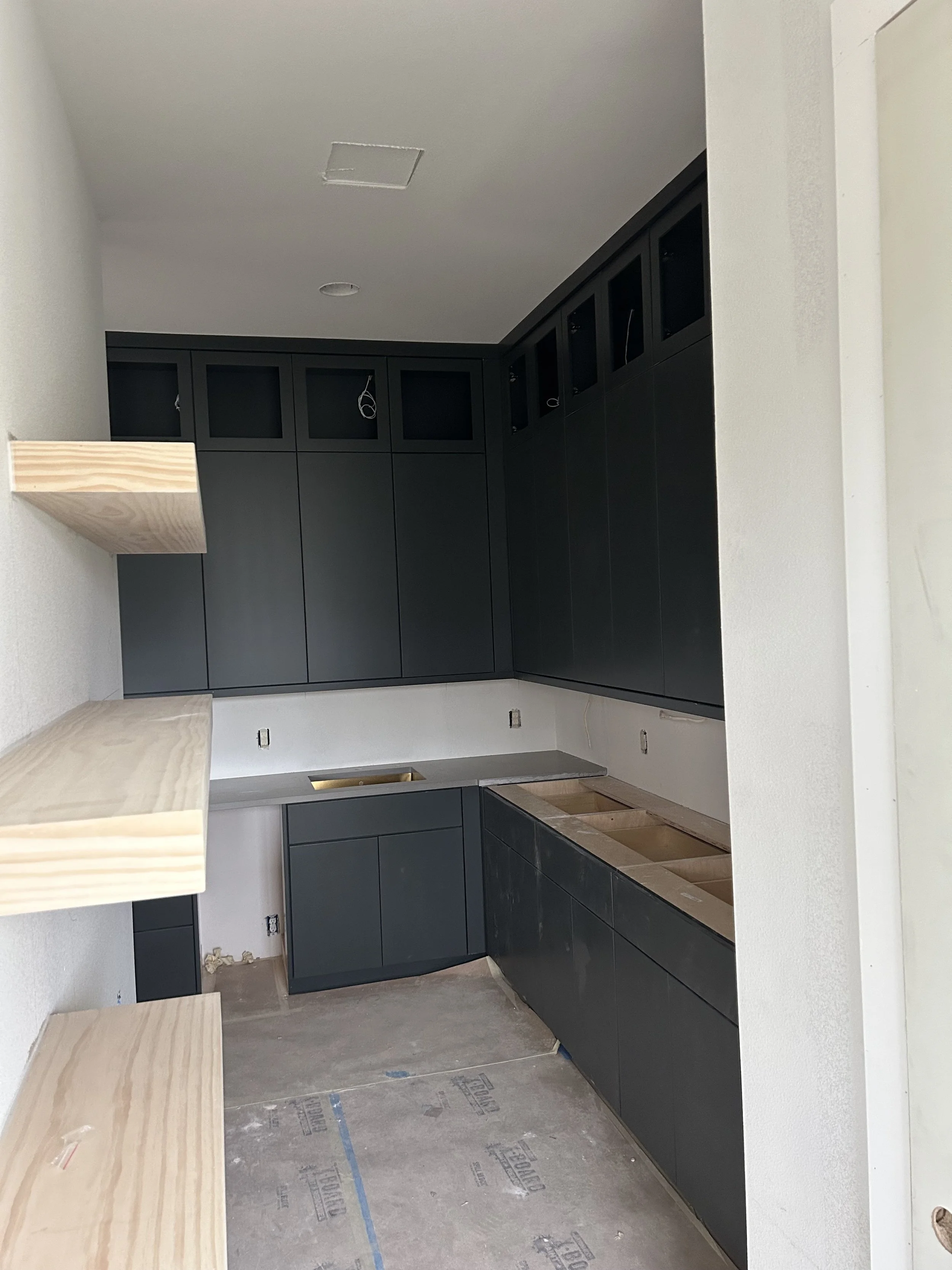 Kitchen under construction with dark cabinets, unfinished countertops, and open shelving on the wall.