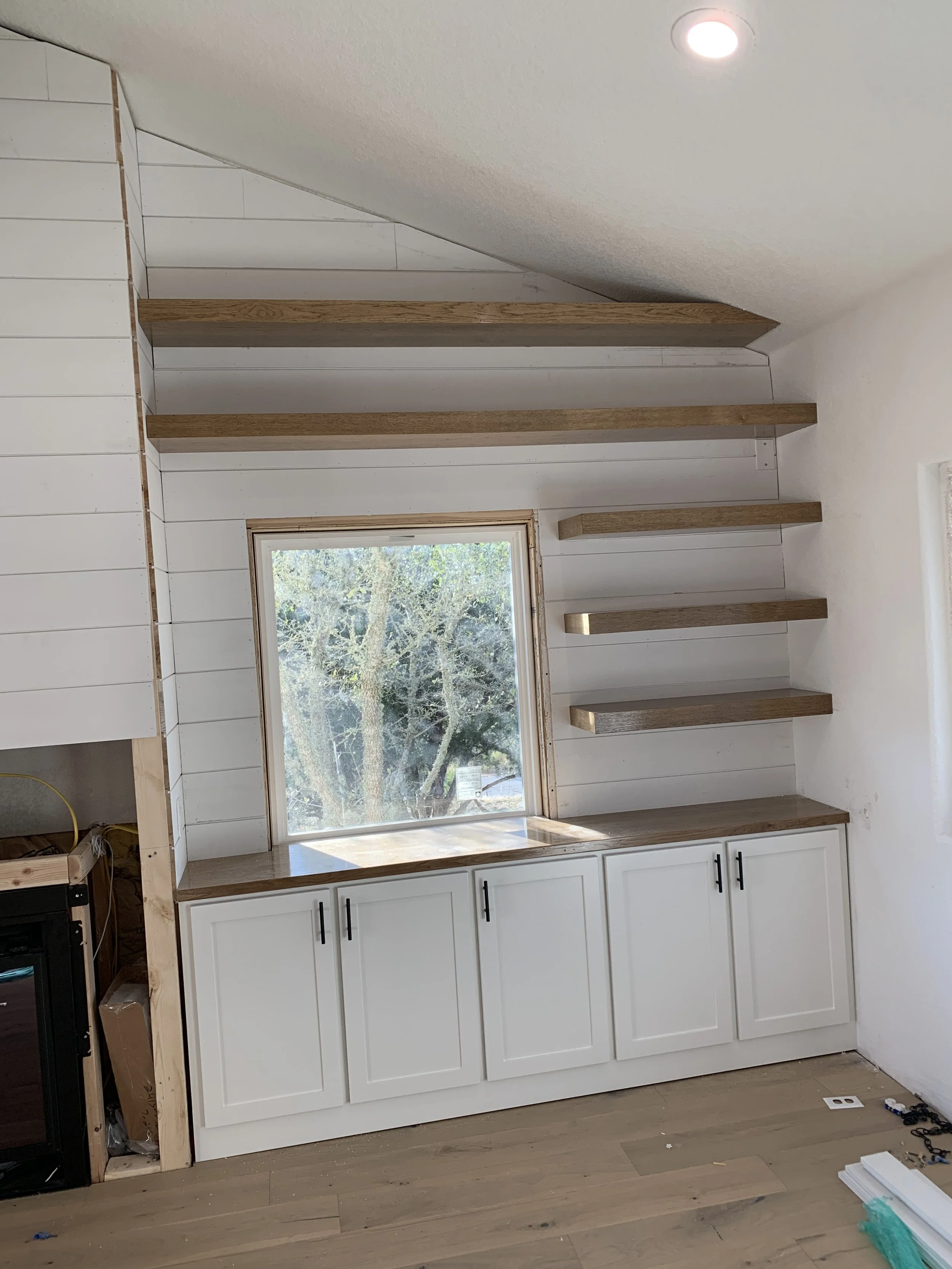 Interior of a room with white cabinets, a wooden countertop, and wooden shelves above a window, natural light coming through the window.