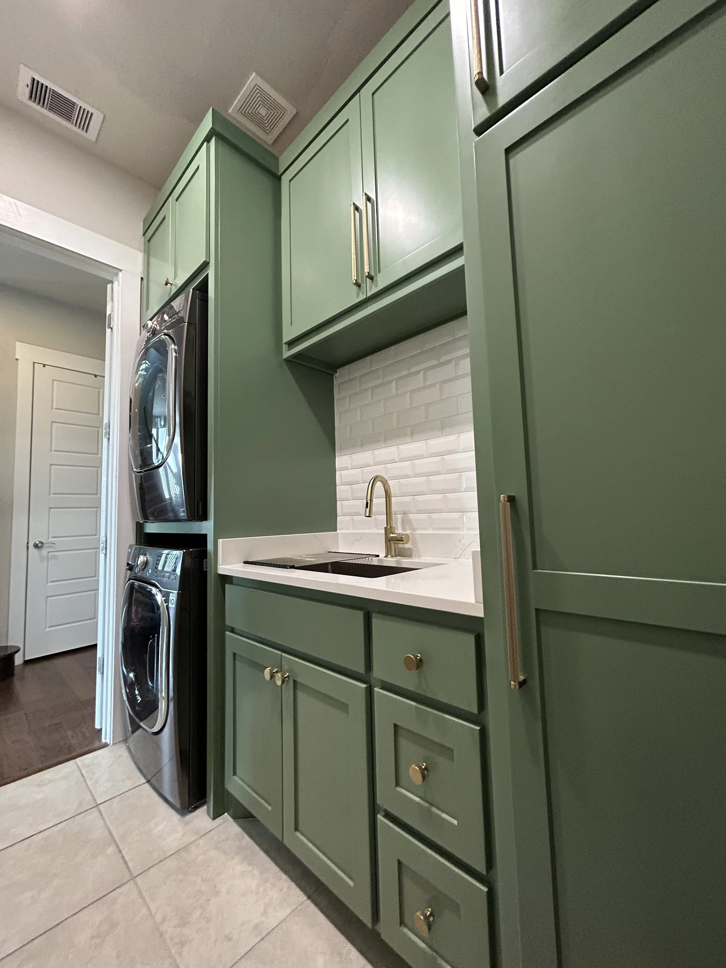 Green kitchen cabinets with gold knobs, a white countertop with a gold sink and faucet, a white brick backsplash, and stacked black washer and dryer units.