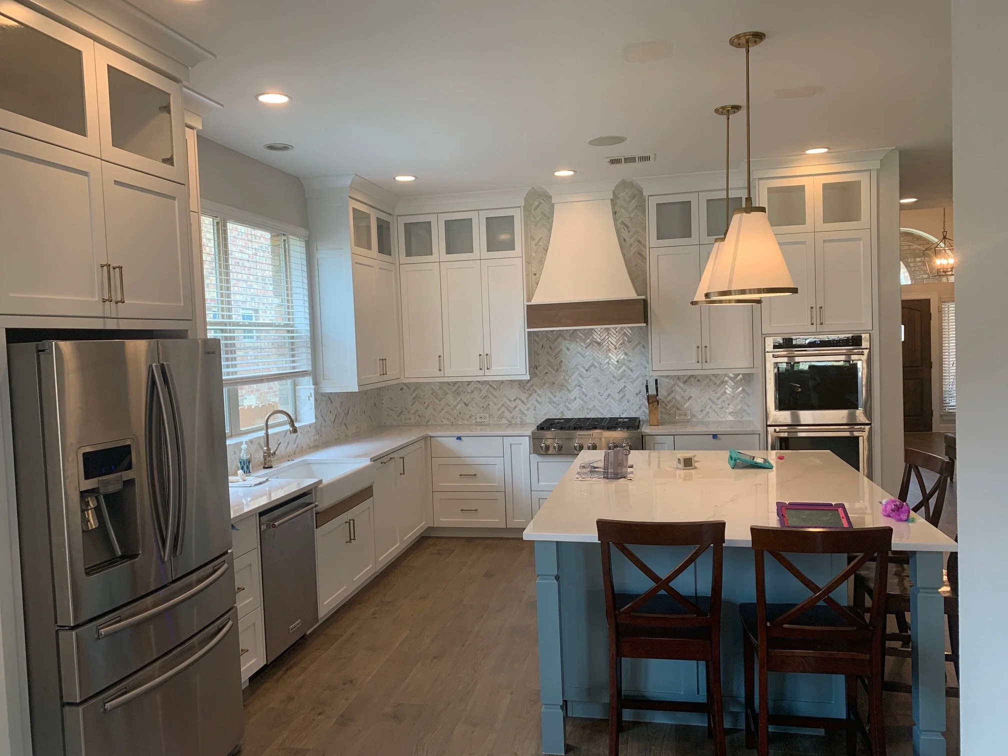 Modern white kitchen with stainless steel appliances, a large island, pendant lighting, and a chevron tile backsplash.