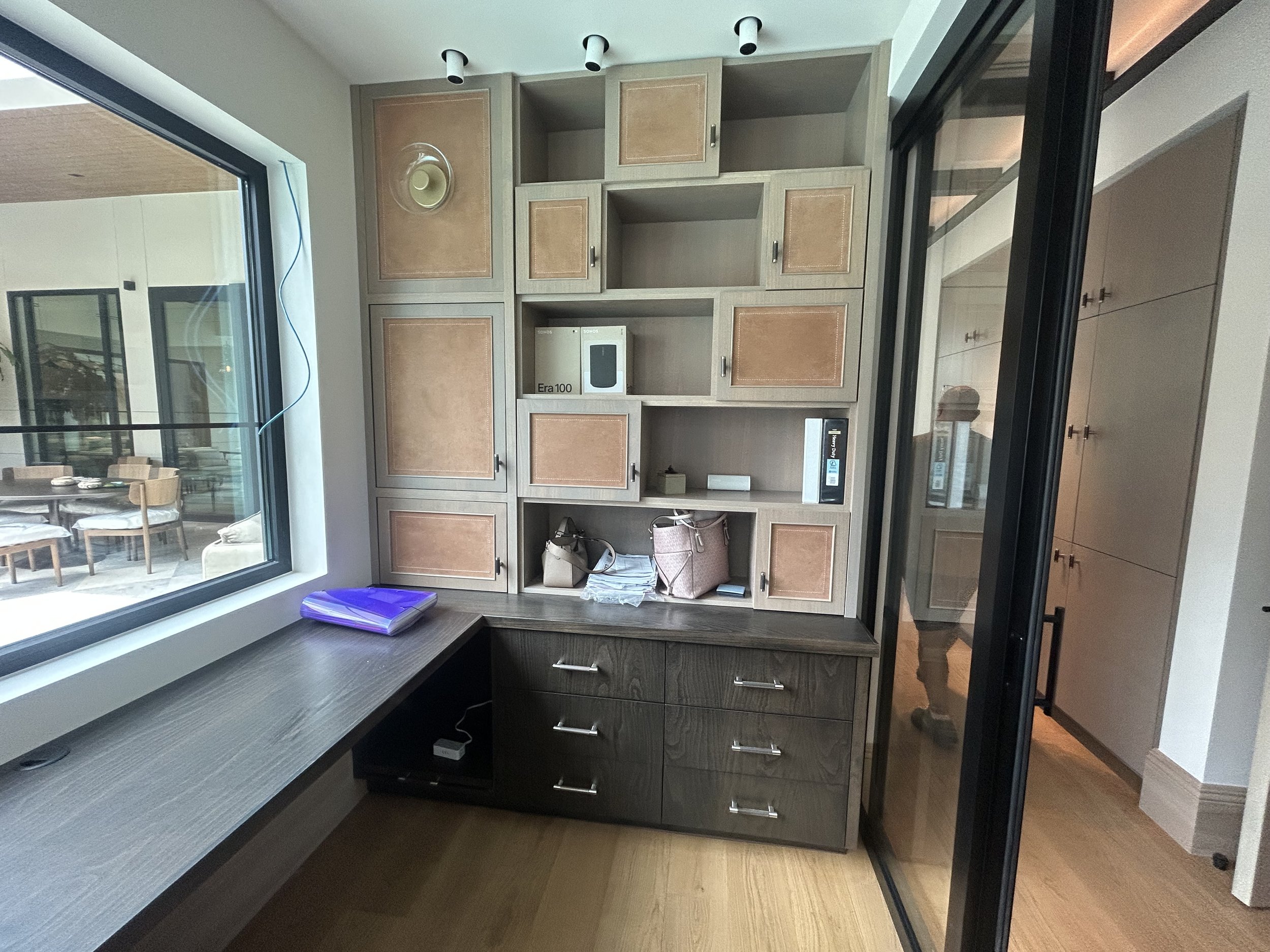 A corner of a room showing a large window, a desk with a lamp, and a built-in wall cabinet with closed and open storage spaces, some containing boxes, books, and handbags. There is a reflection of a person taking the photo in a glass door.