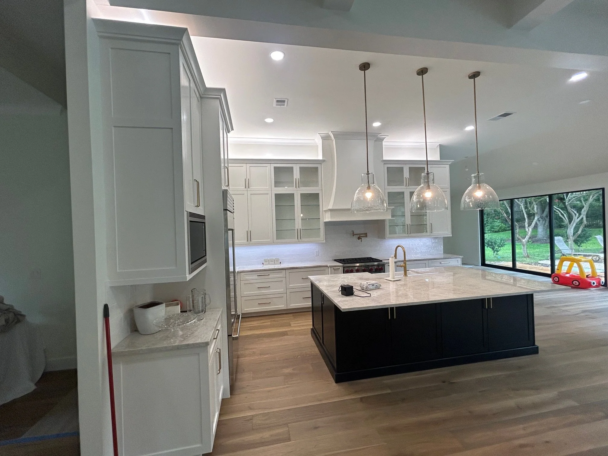 Modern kitchen with white cabinets, a large center island with a marble countertop, three hanging pendant lights, stainless steel appliances, and a large window showing a backyard with trees and outdoor chairs.