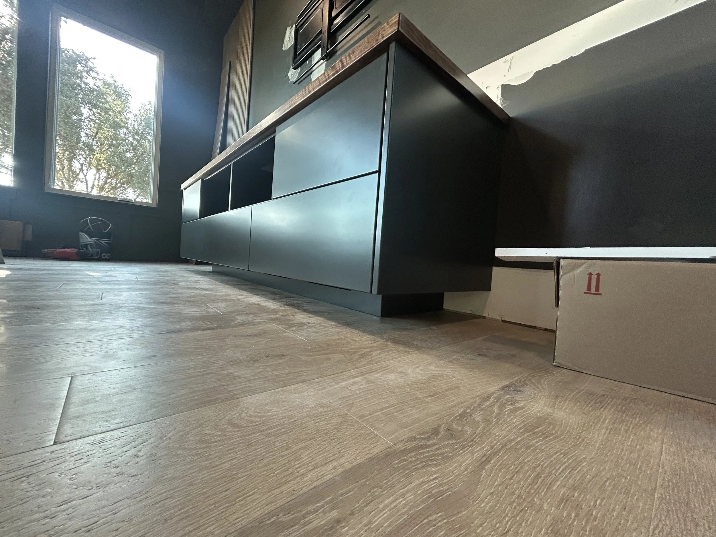A kitchen with a black cabinet, a window and a wooden floor. There is a cardboard box on the floor near the wall.