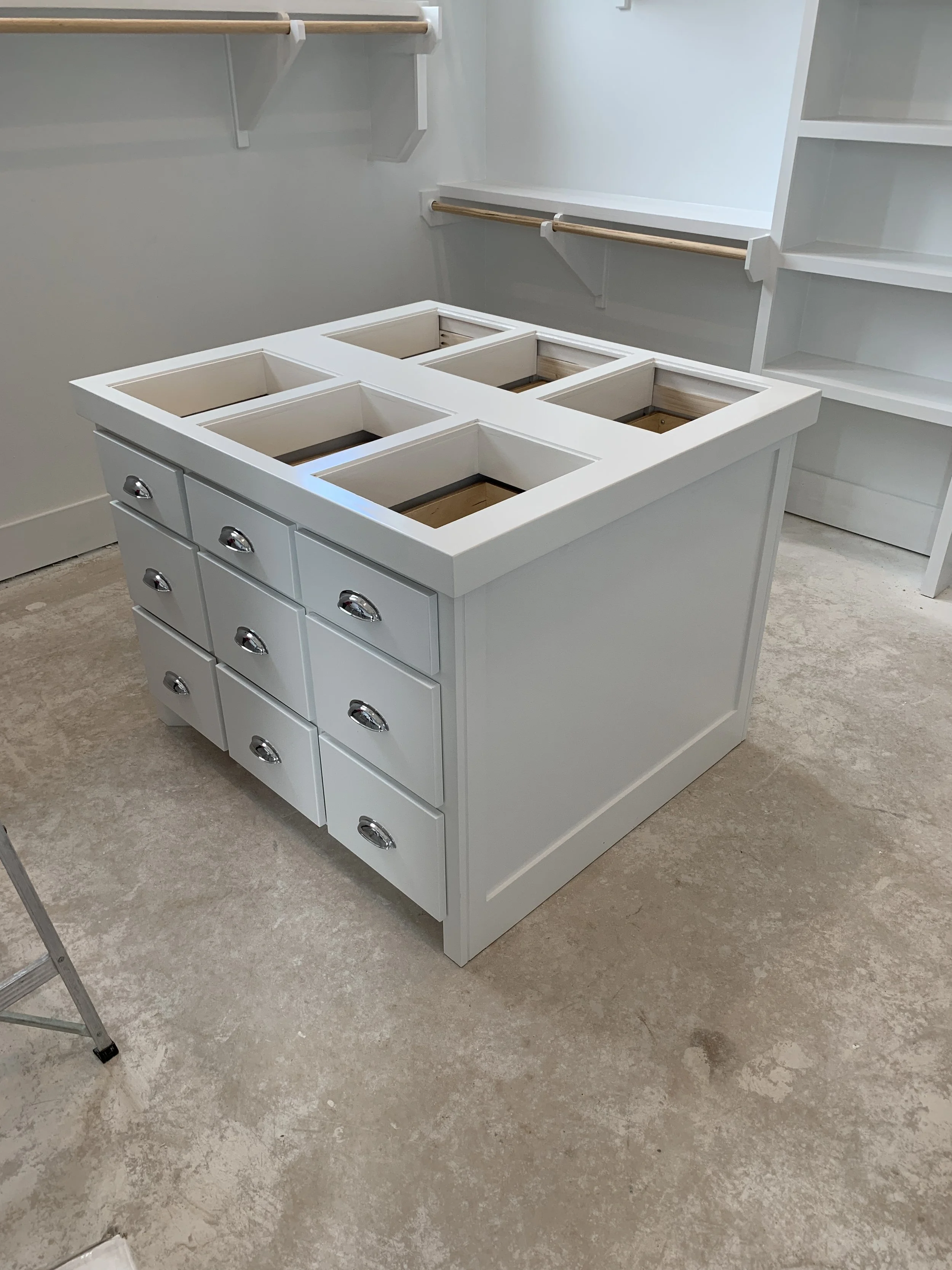 A white rectangular kitchen island with multiple drawers and open cutouts on top, situated on a beige carpeted floor.