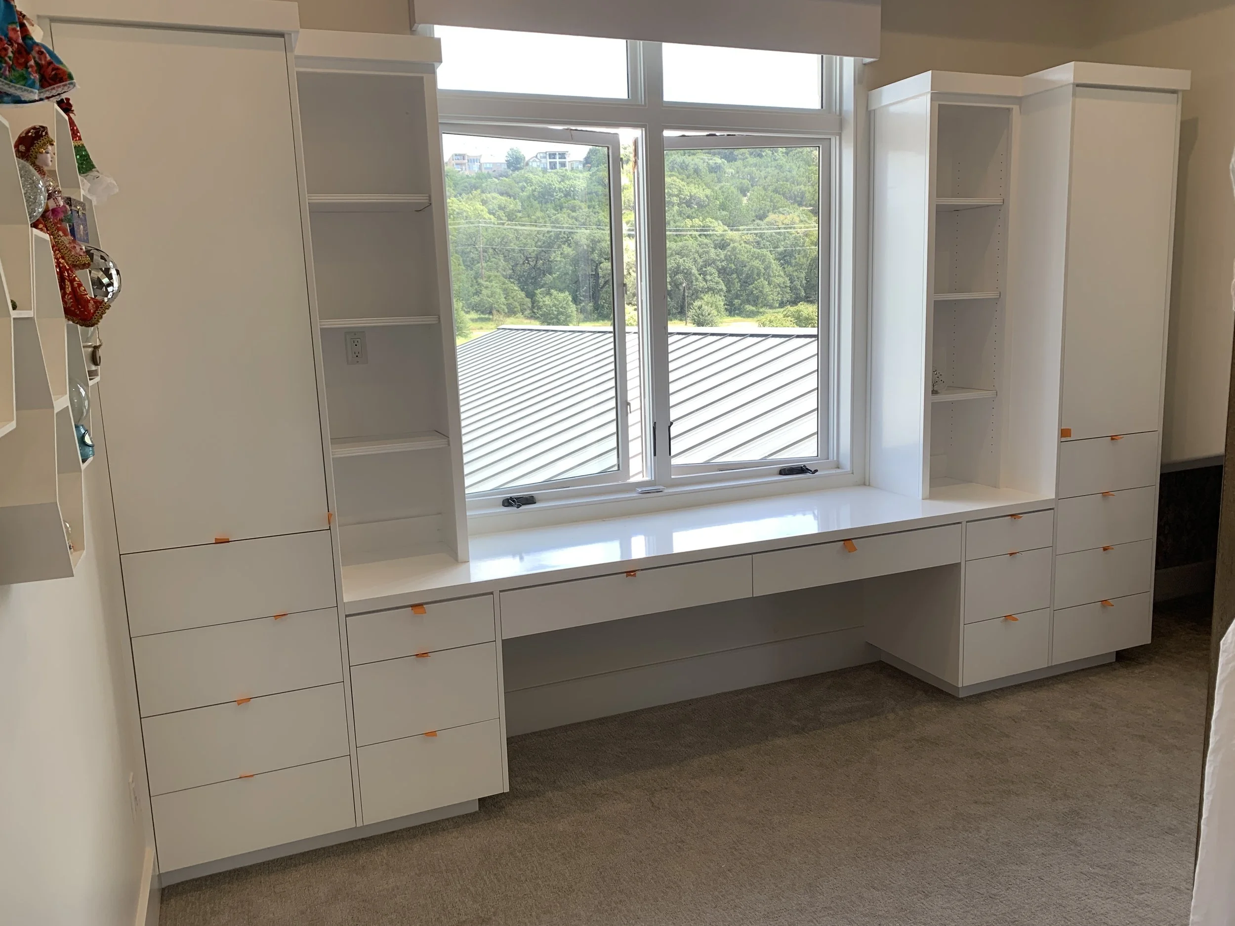 Empty white built-in desk and shelving unit under a window in a room, with some small orange markers on the drawers and shelves.