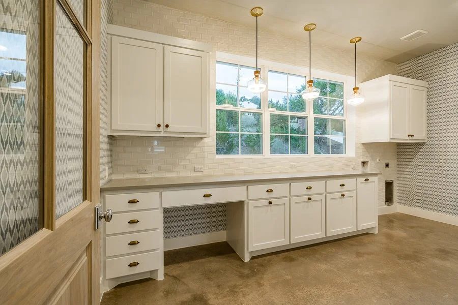 Empty kitchen with white cabinets, a countertop, natural light from windows, pendant lights, and patterned wallpaper on one wall.