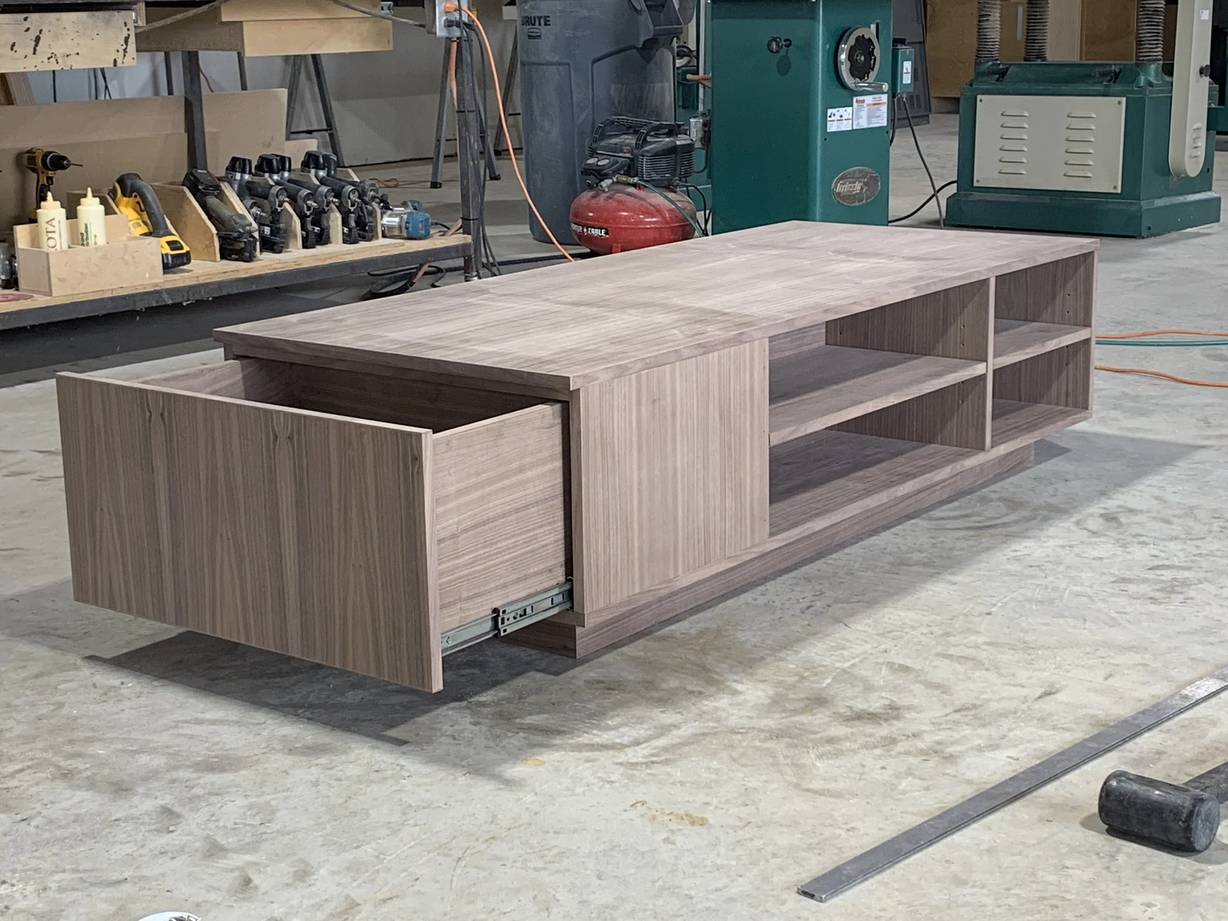 Wooden TV stand with open shelves and a sliding drawer on a workshop floor.