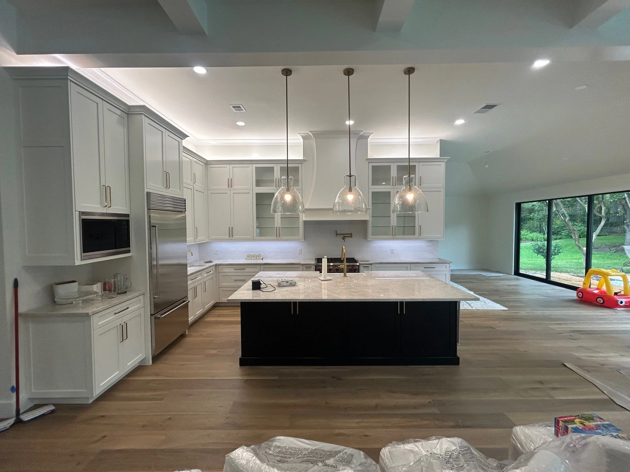 A modern kitchen with white cabinetry, a large center island with black base and white countertop, three pendant lights hanging above, stainless steel appliances, wood flooring, and large sliding glass doors leading to an outdoor yard with a swing se
