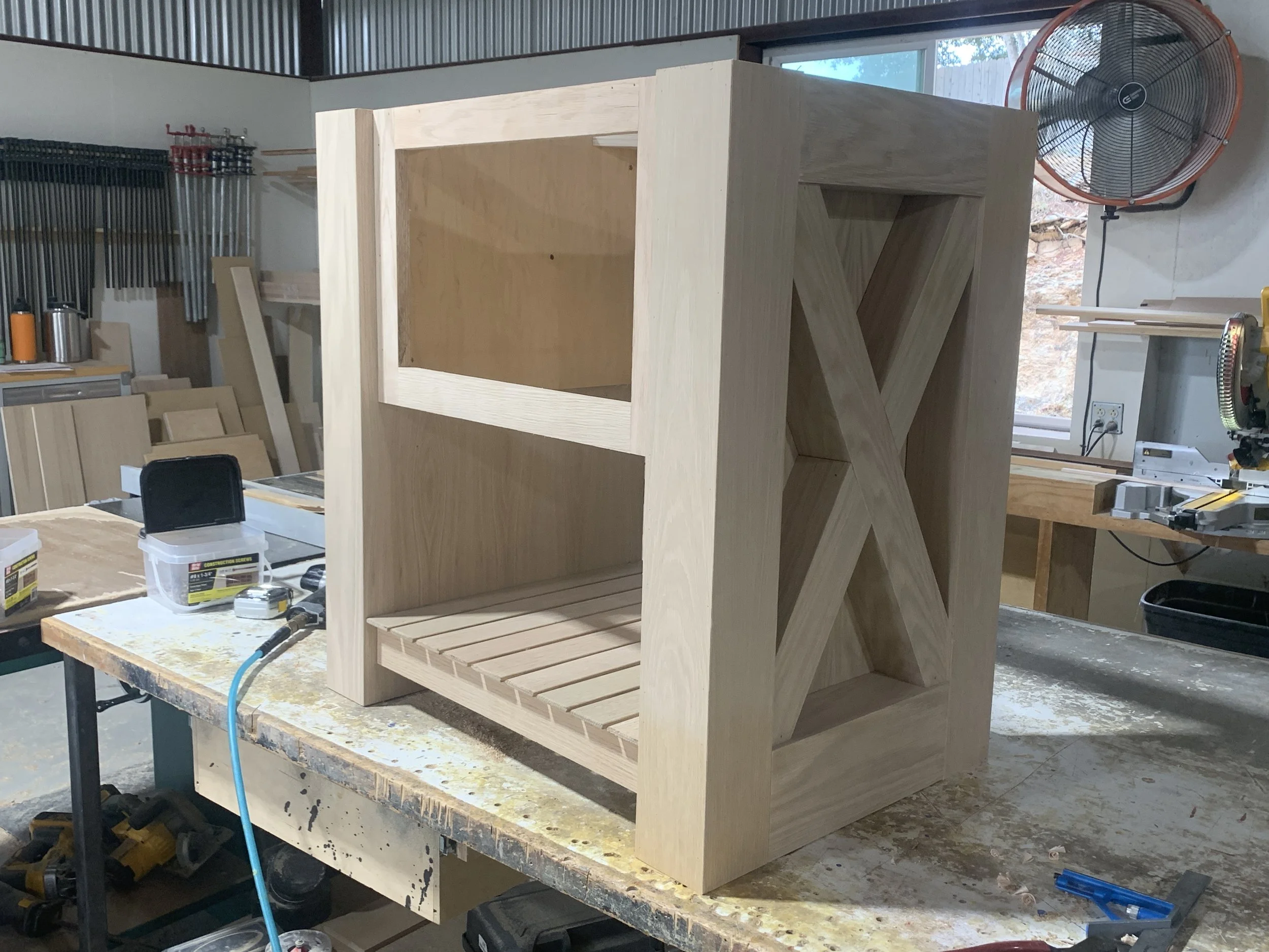 Wooden furniture piece under construction in a woodworking workshop, with a large fan in the background.