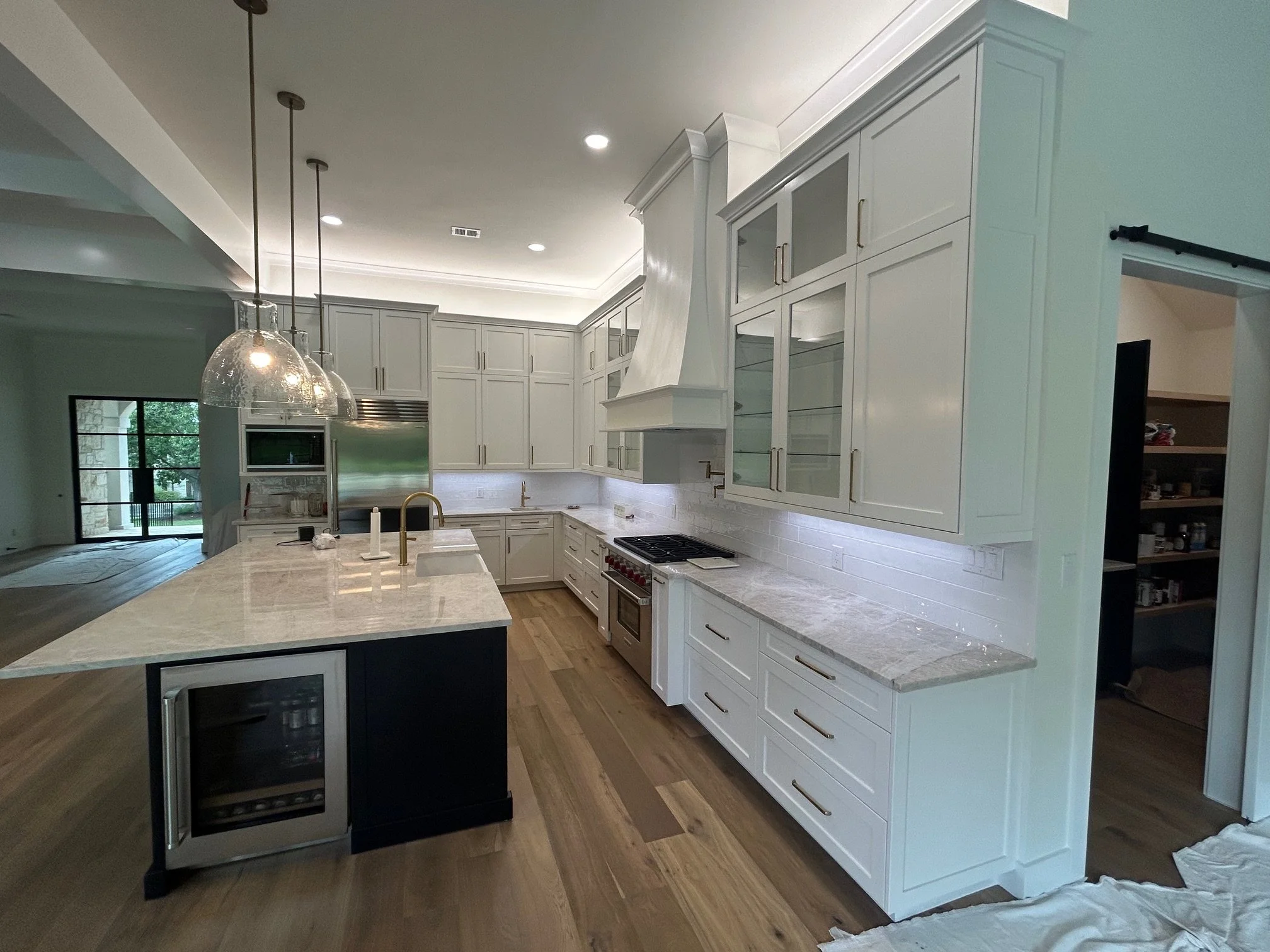 Modern kitchen with white cabinets, marble countertops, a central island with a wine fridge, gold fixtures, and pendant lighting, with a view of a living area and sliding glass door.