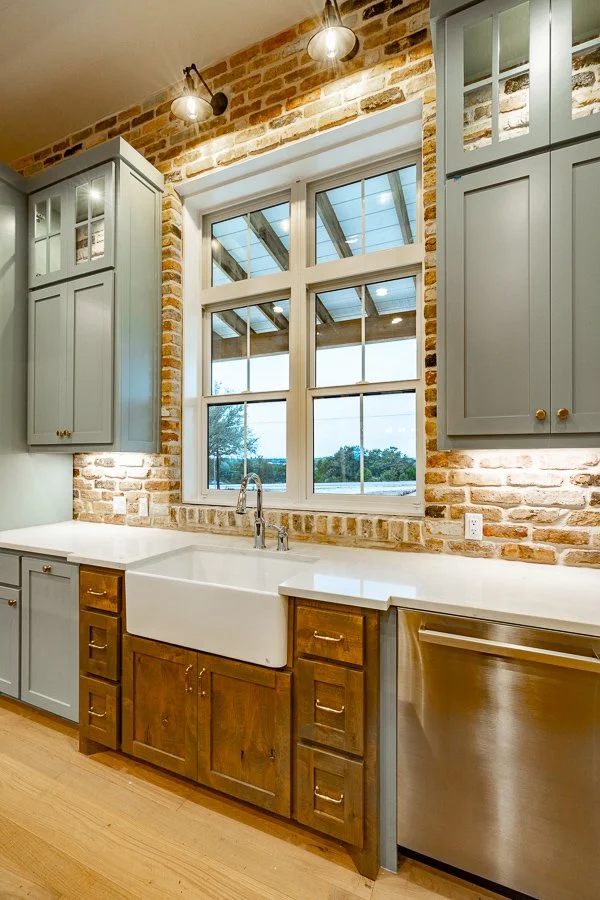Bright kitchen with exposed brick wall, large window, farmhouse sink, light blue upper cabinets, wooden lower cabinets, and stainless steel dishwasher. Overhead lighting fixture.