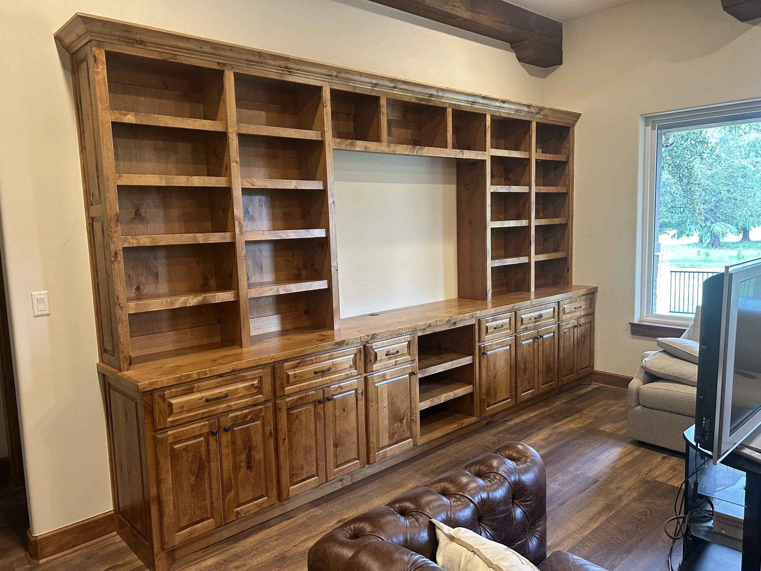 Large wooden bookcase with open shelves and cabinets, located in a living room with a window, beige walls, and hardwood floors.