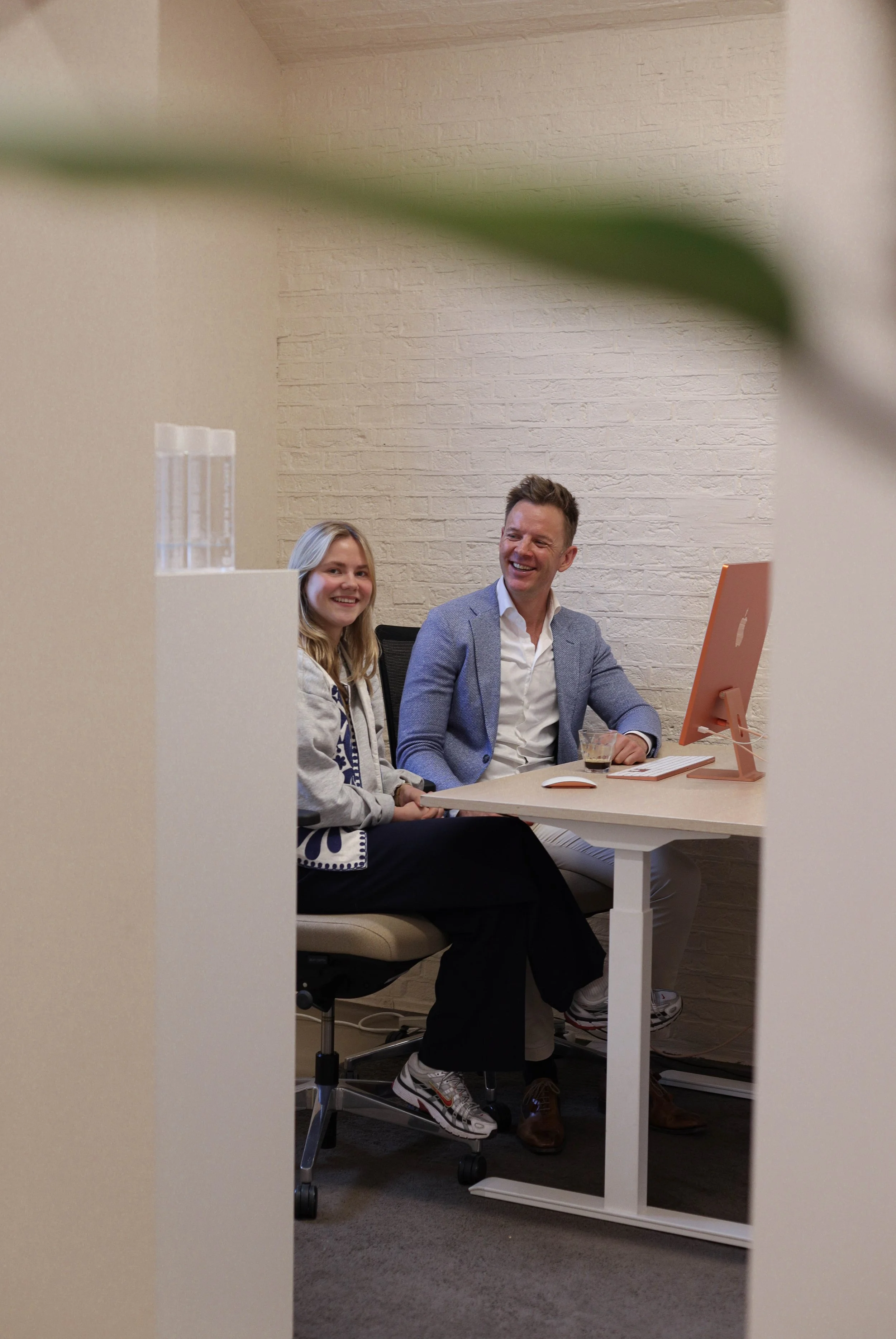 To personer sitter ved et kontorbord og ler, sett gjennom en åpen dør.