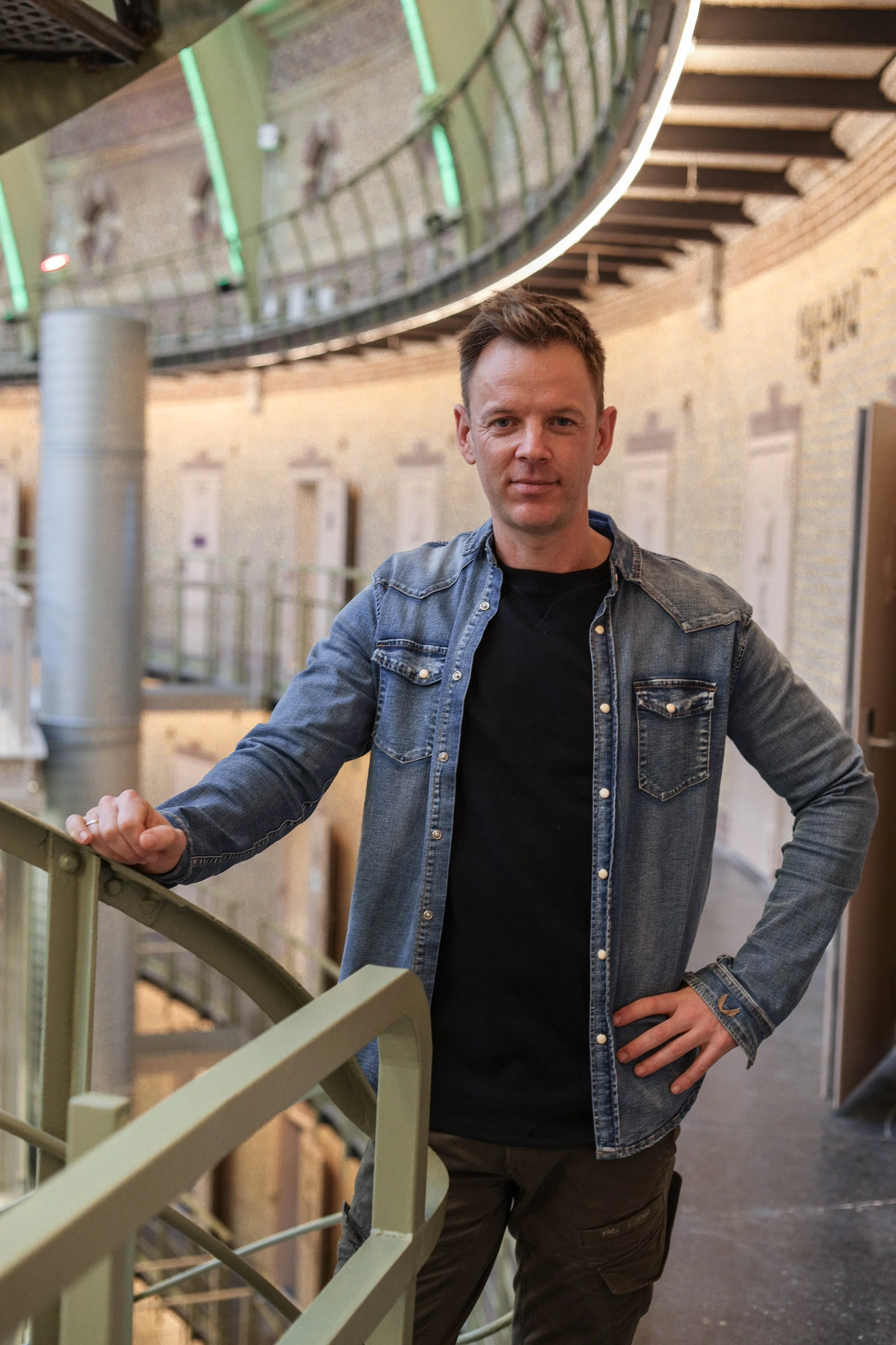 En mann står ved en gelender i et indendørs industrimiljø, iført en blå denimjakke og svart t-skjorte.