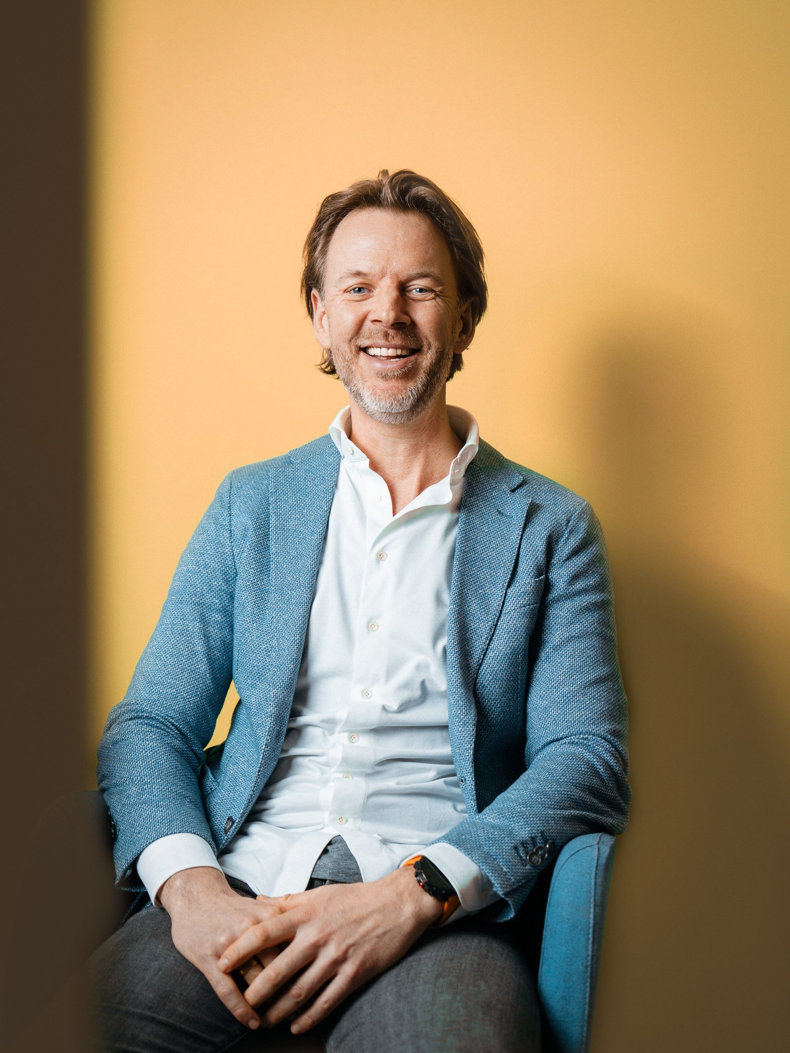 Et portrett av en smilende mann med brunt, litt krøllet hår og skjegg, iført blazer og hvit skjorte, sittende mot en gul bakgrunn.