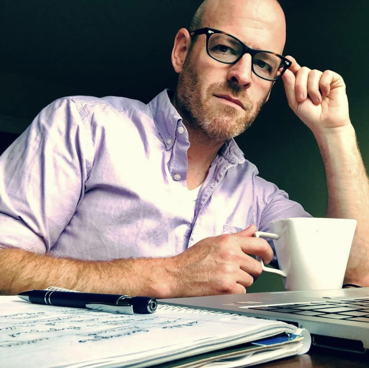 A man with glasses, a beard, and a light purple shirt sitting at a desk with a notebook, pen, coffee mug, and a laptop.